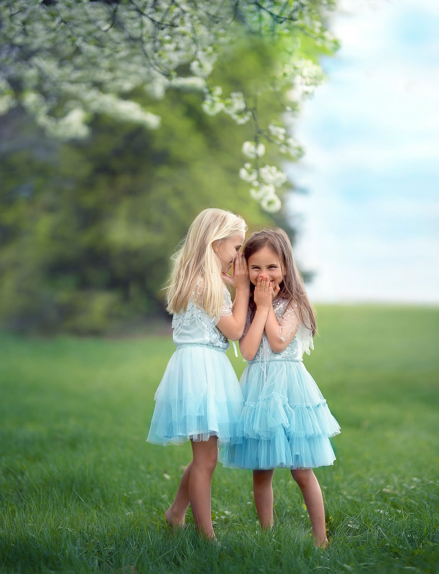 Two young friends in matching blue Tutu Du Monde dresses whisper and giggle together in a sunlit spring meadow.
