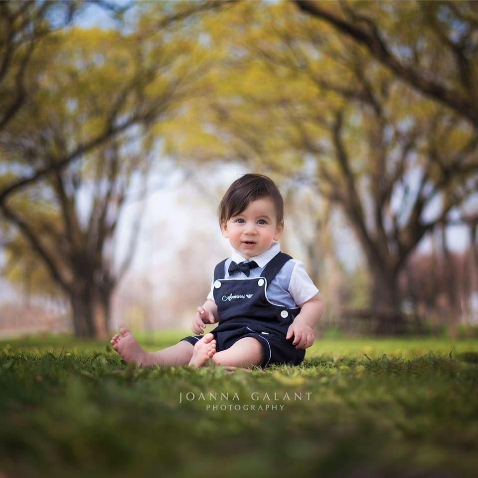 A small child sits in overalls on grass beneath yellow autumn trees in a soft focus portrait.