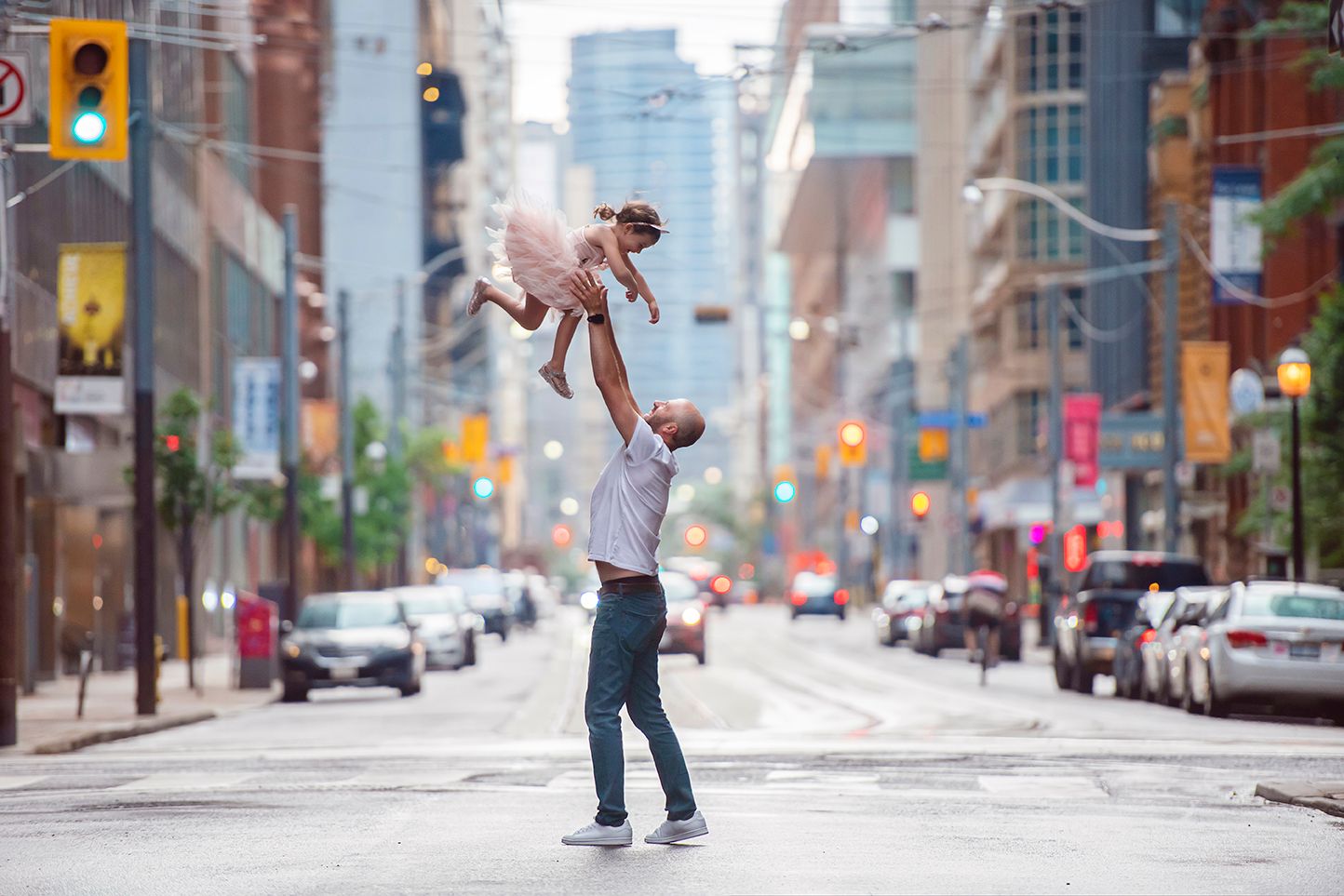 Dad lifts child playfully high in the air on empty city street with tall buildings and parked cars in Toronto.