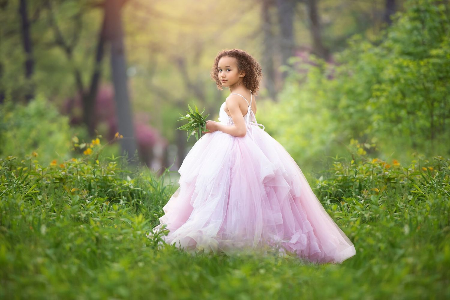 A child in a flowing pink Anna Triant Couture dress twirls joyfully in a dreamy forest setting surrounded by green foliage.