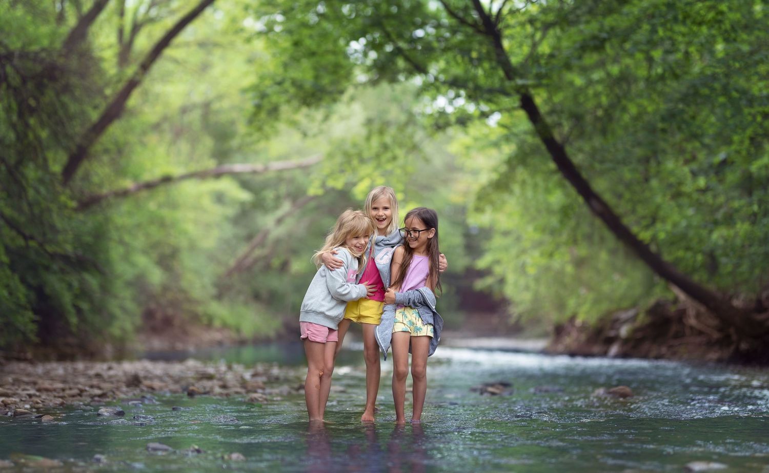 There friends children stand together in a flowing stream surrounded by lush green trees on a sunny summer day.