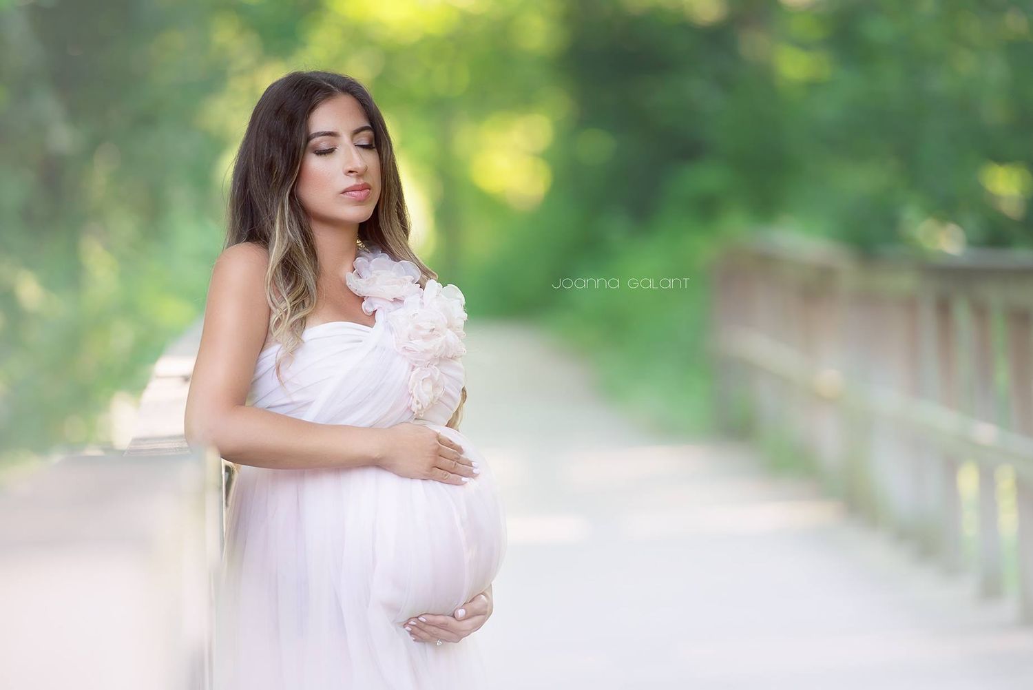 A pregnant and expecting  mom  in a flowing white dress poses on a bridge surrounded by dreamy bokeh and soft natural light.
