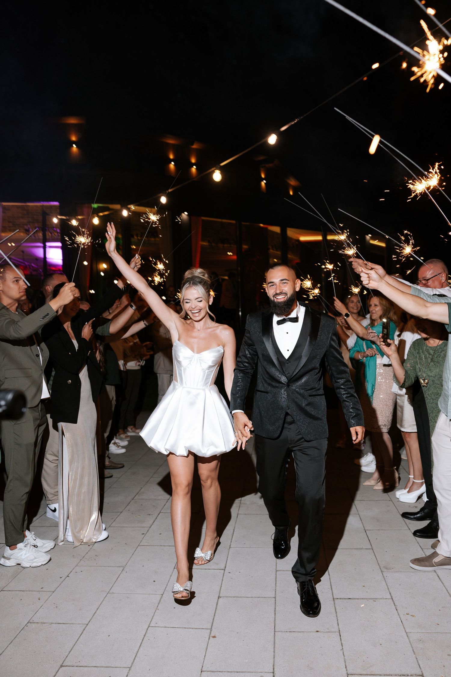 A couple exits their wedding reception under sparklers while the bride wears a short white dress and groom in black tuxedo.