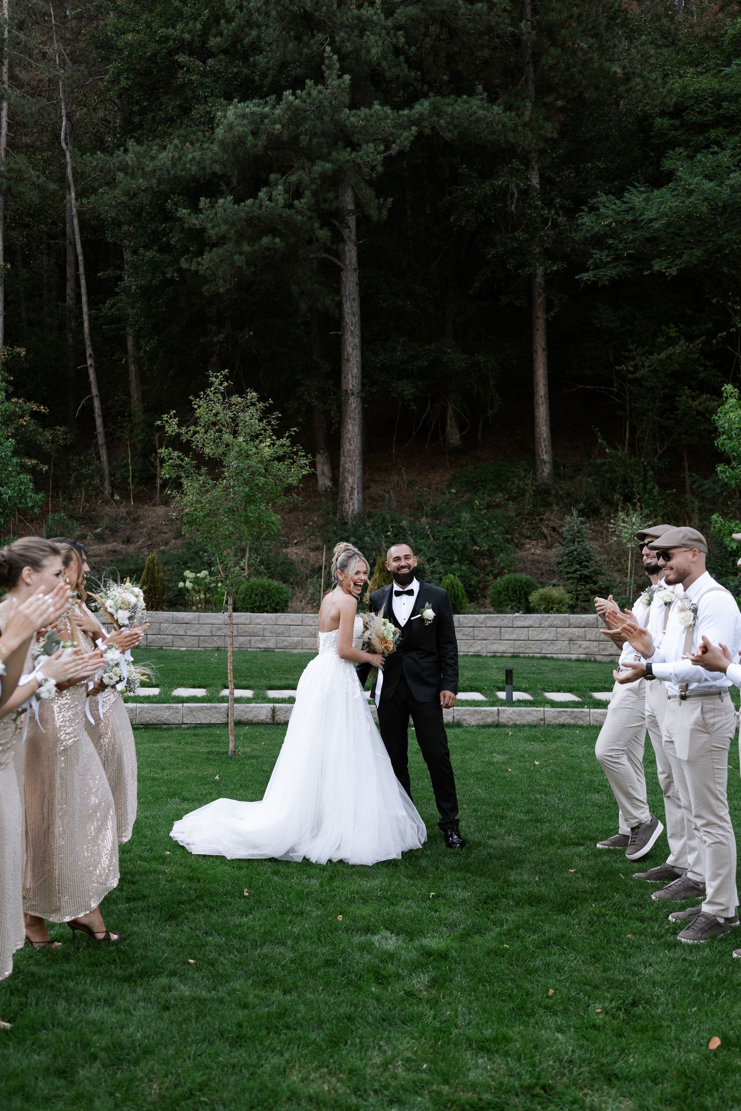 Wedding party members play tug of war on a grassy lawn with a beautiful forest backdrop during outdoor reception.