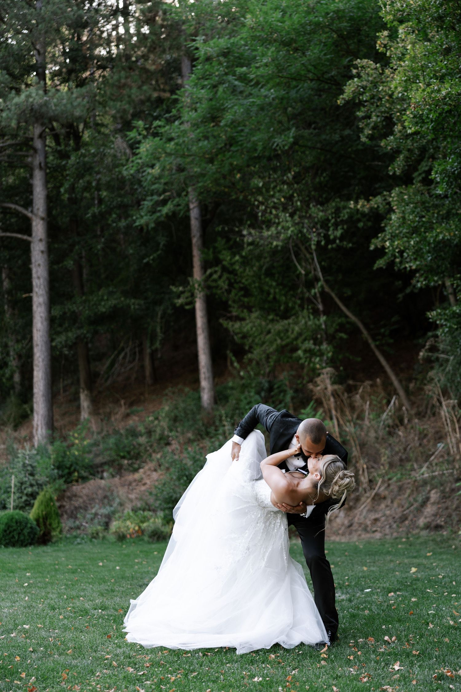 A couple in wedding attire shares romantic moments in a lush garden setting with trees in the background.