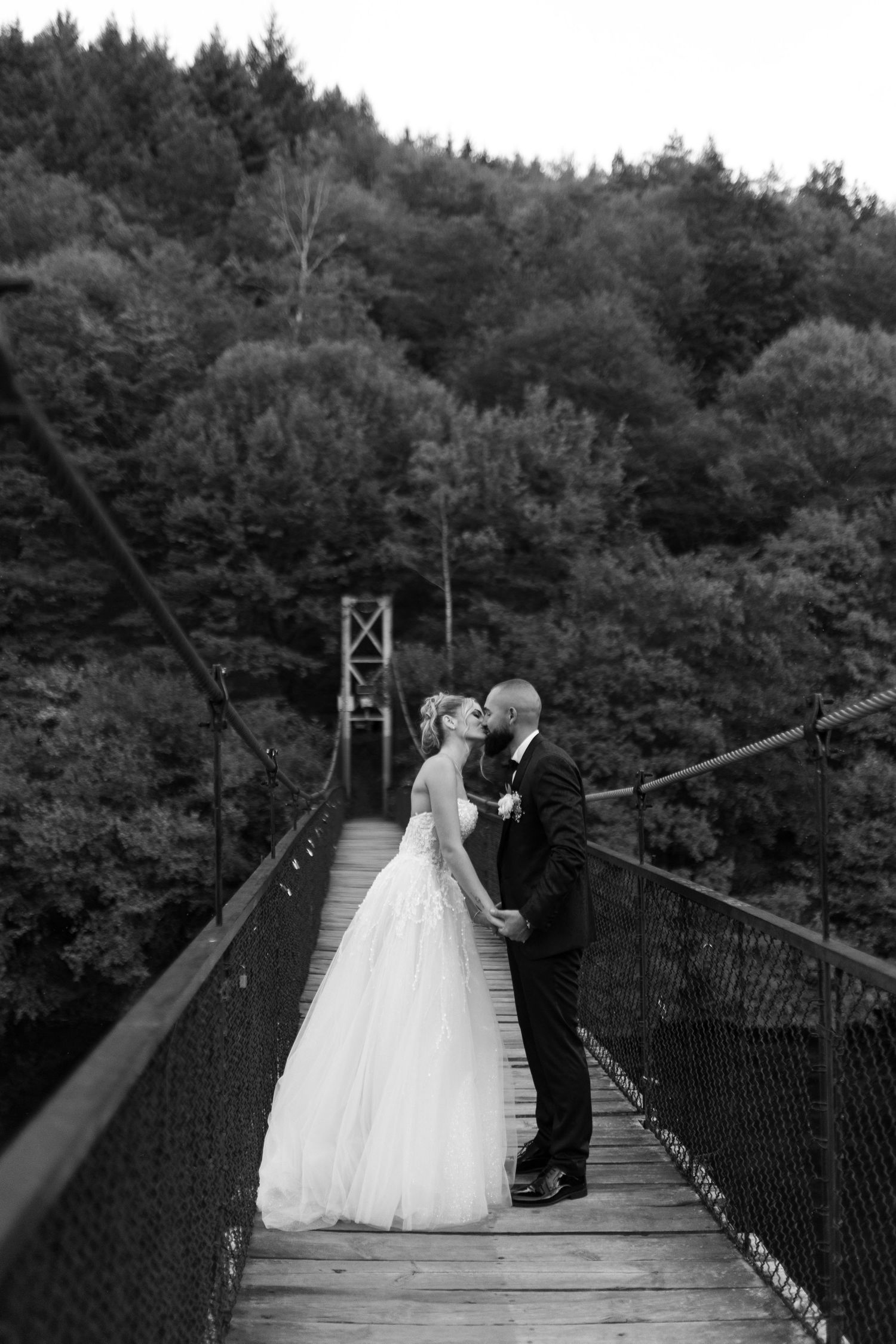 A couple shares an intimate moment on a rustic suspension bridge surrounded by lush forest scenery.