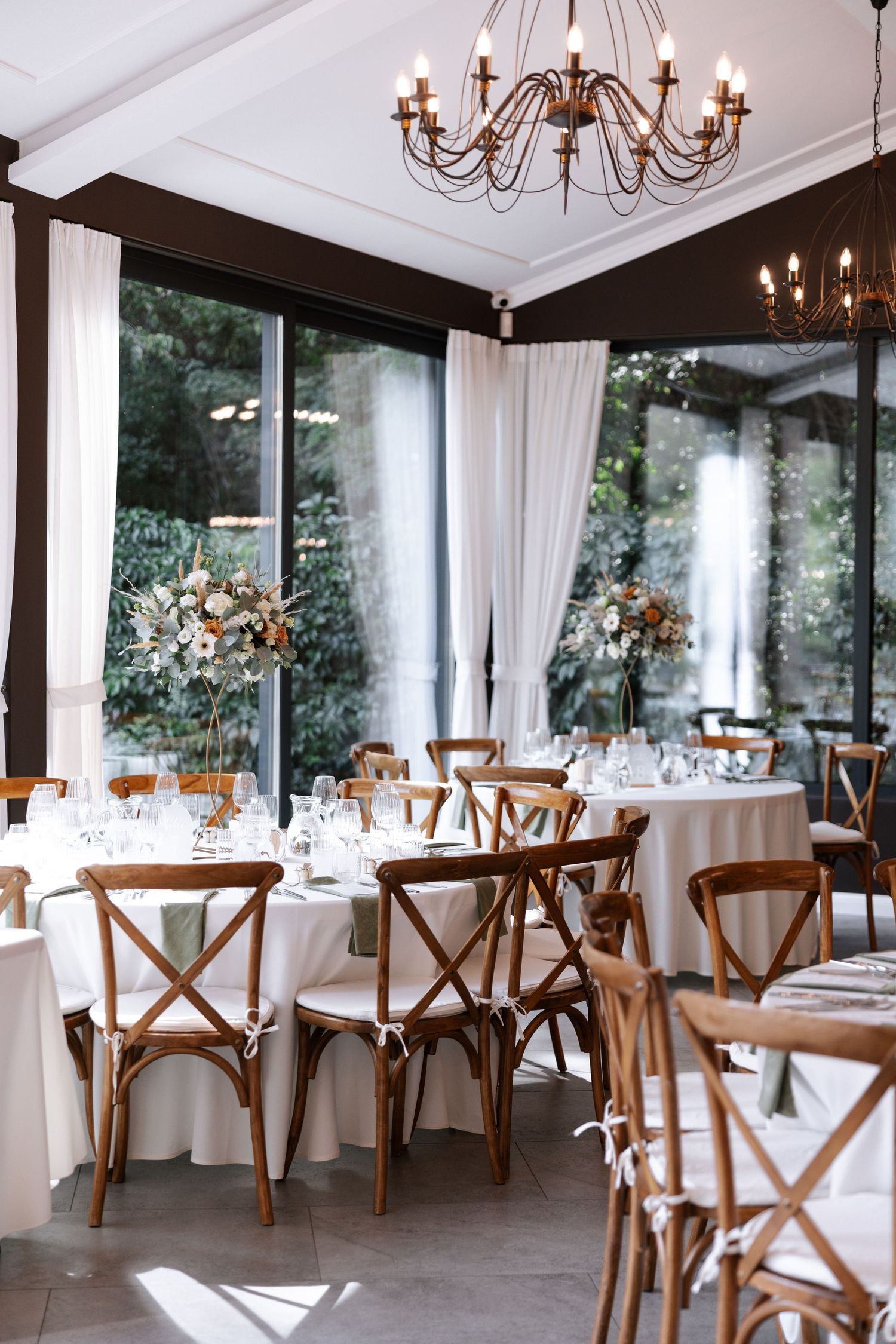 Elegant wedding reception venue with white tablecloths, wooden cross-back chairs, and a vintage chandelier in natural light.