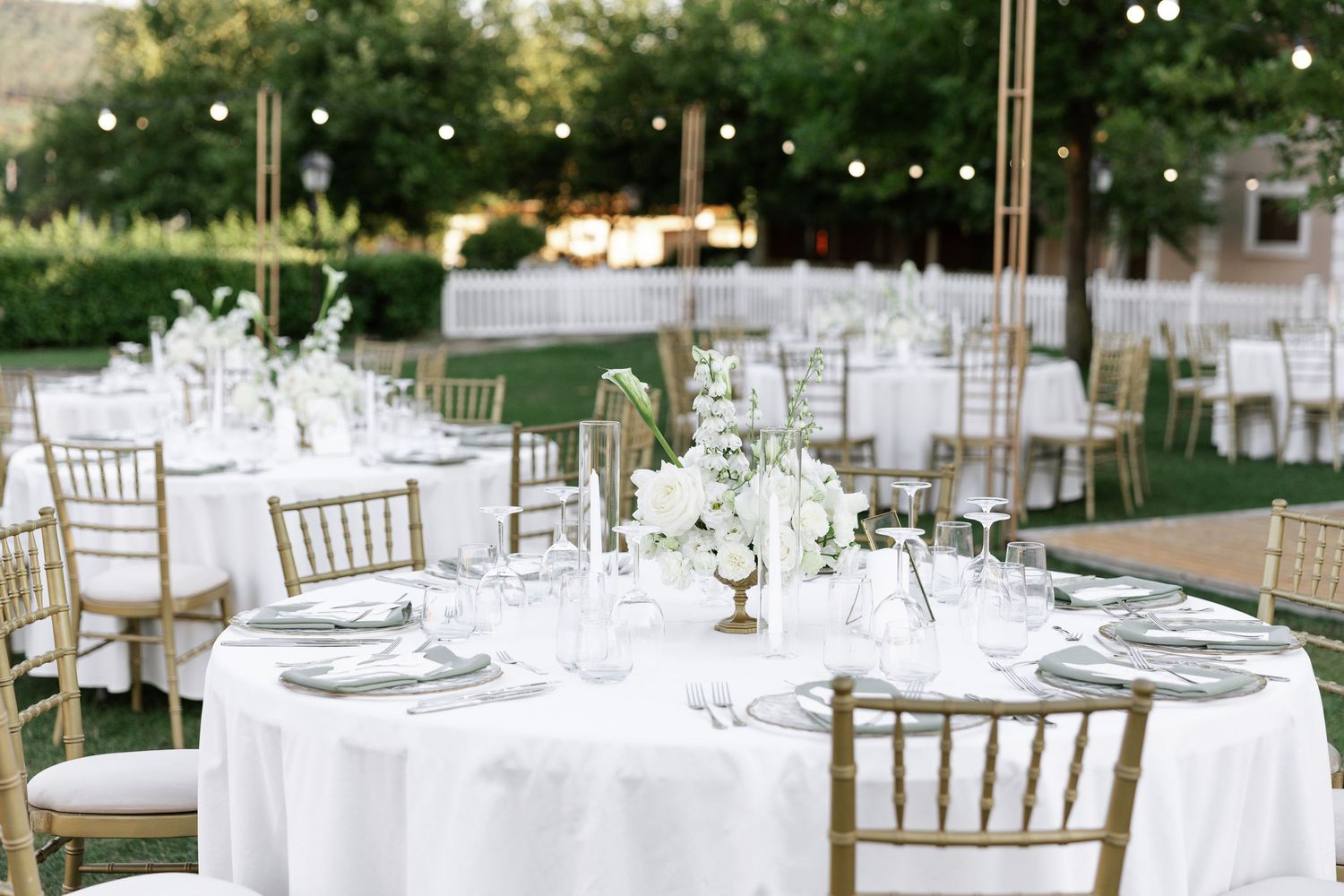 Elegant outdoor wedding reception setup with white tablecloths, gold chiavari chairs, and floral centerpieces on manicured lawn.