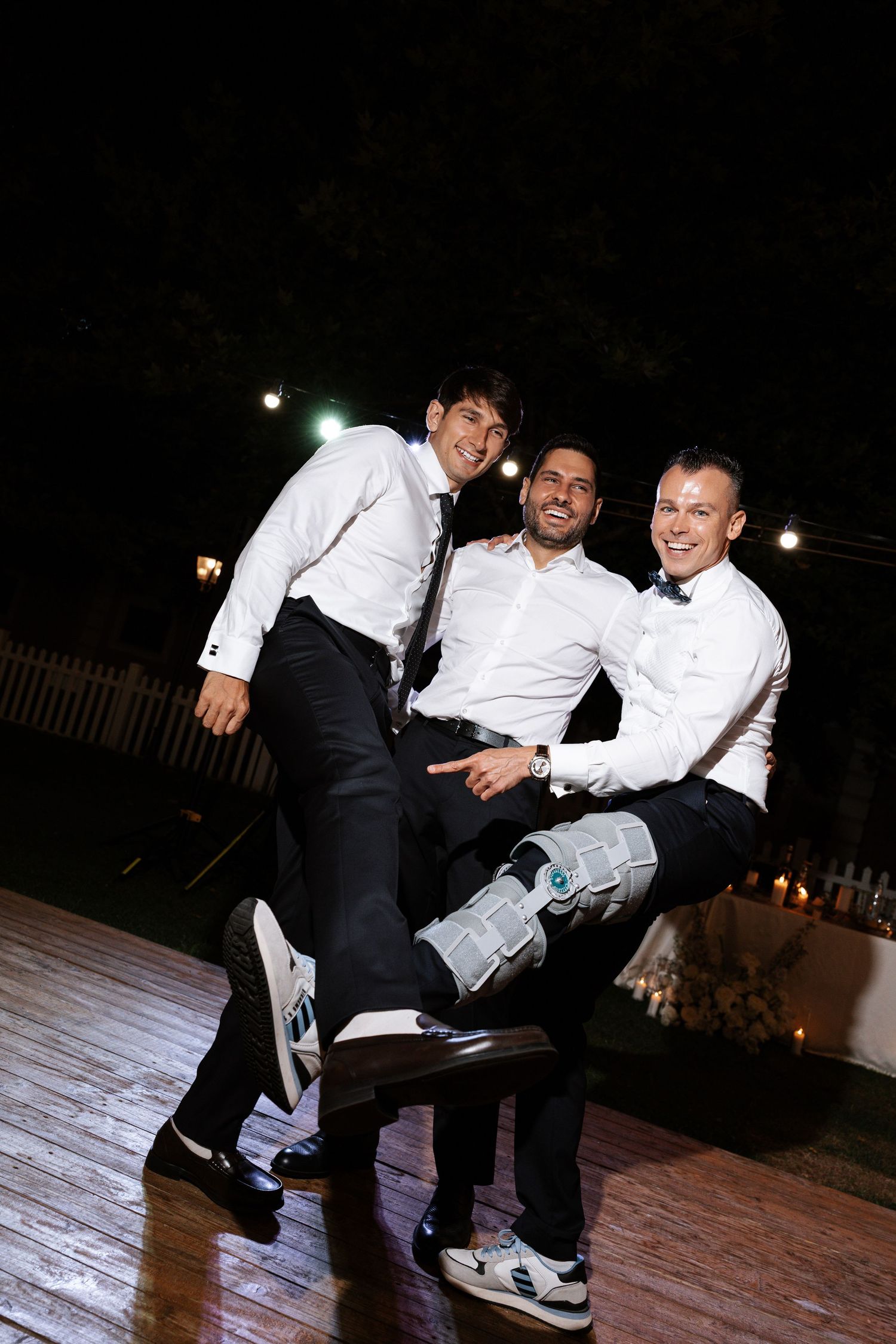 Three people in formal attire laughing and posing together on a wooden dance floor at night.