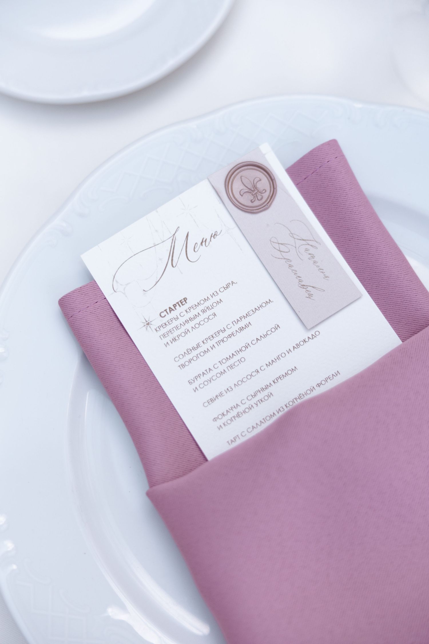 Elegant wedding menu card with wax seal displayed on mauve napkin and white plate.