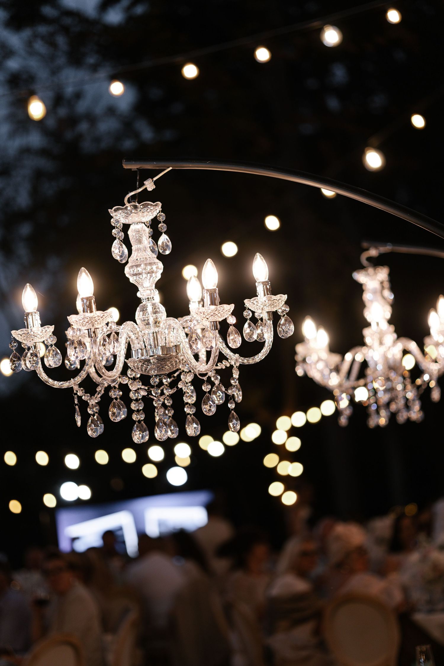 Crystal chandeliers illuminate outdoor evening event with bokeh lights in background.