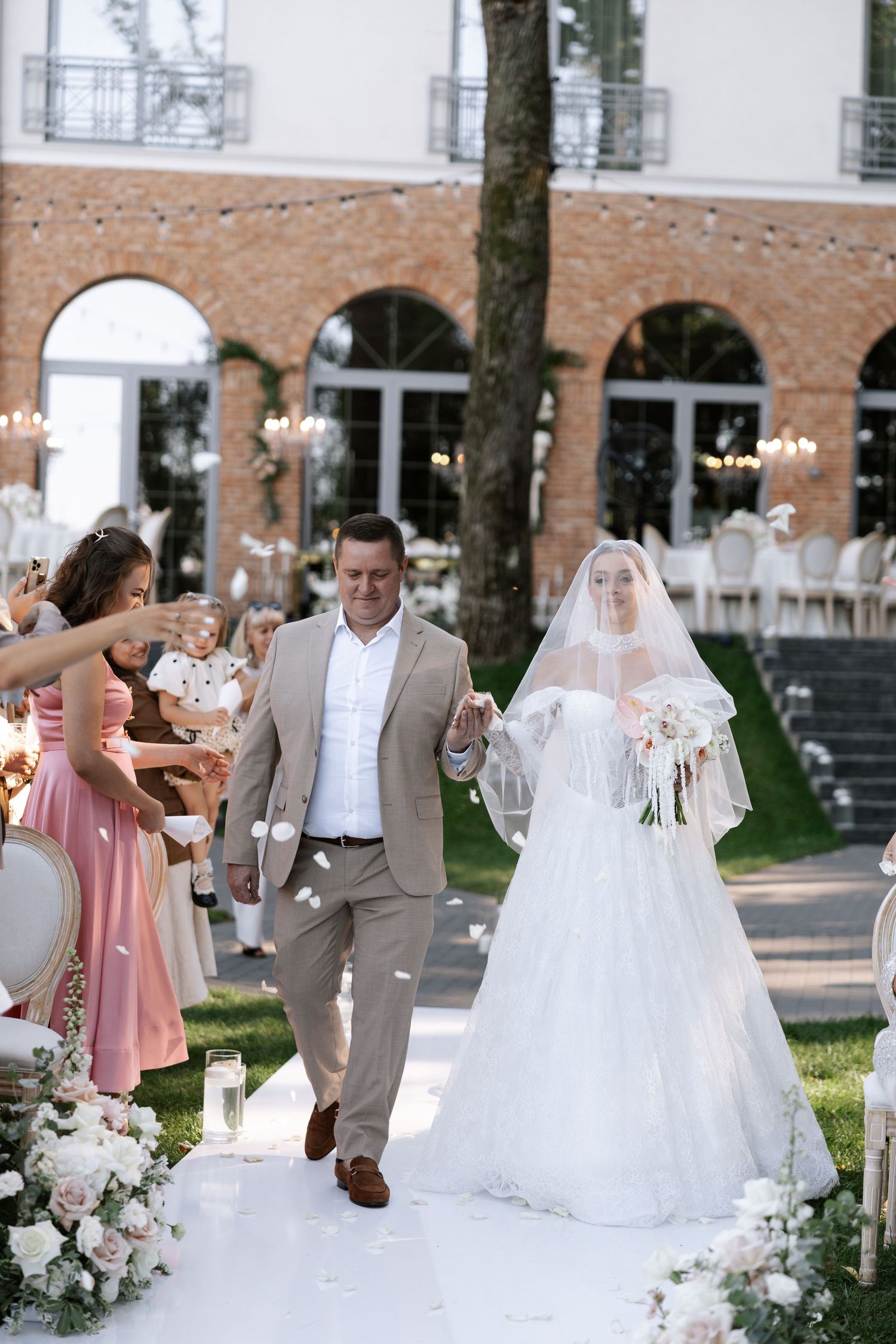 Wedding party in gray suits and pink dresses gathered outdoors near a brick building.