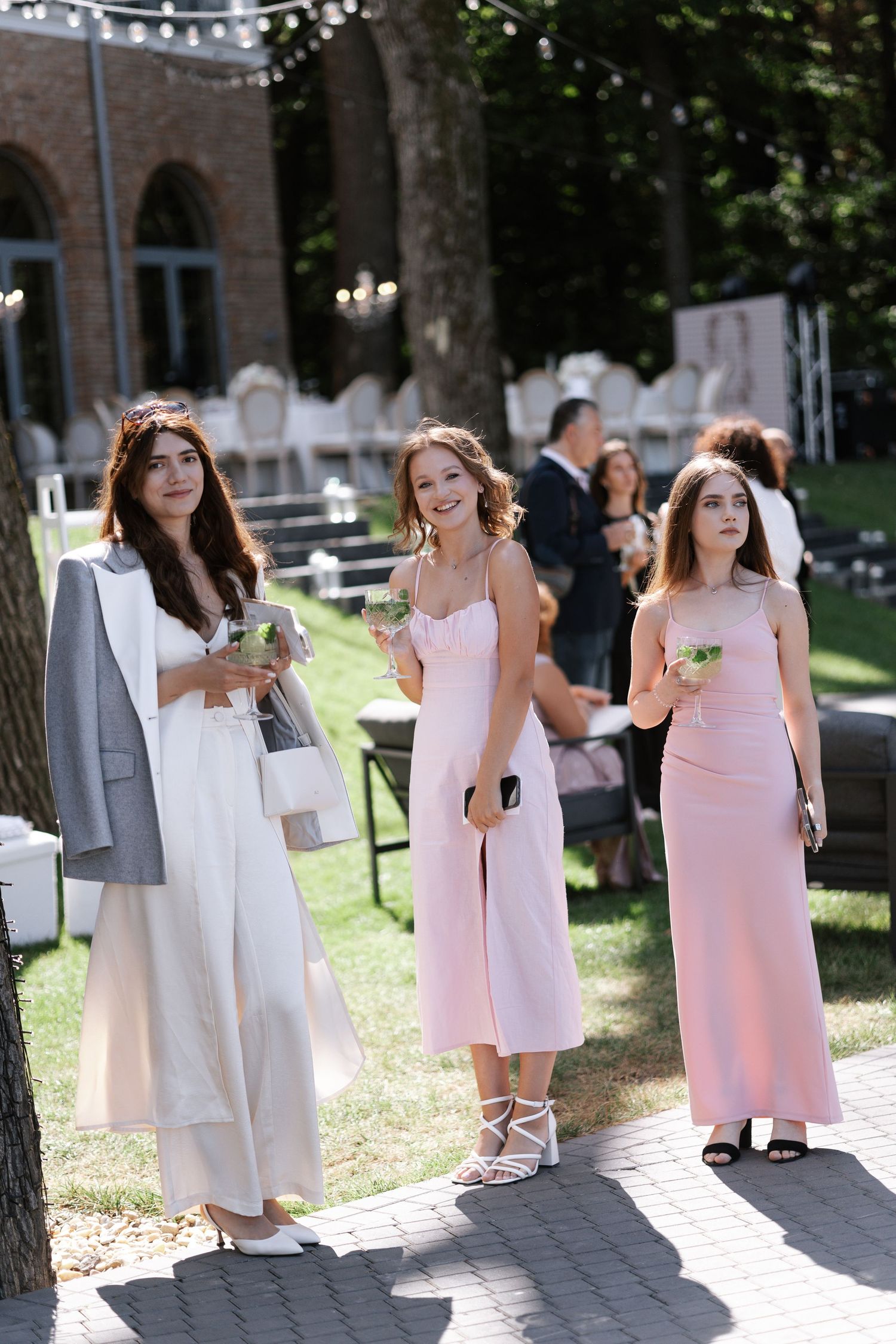 Young bridesmaids in pink and white dresses stand together on stone steps during an outdoor wedding ceremony.