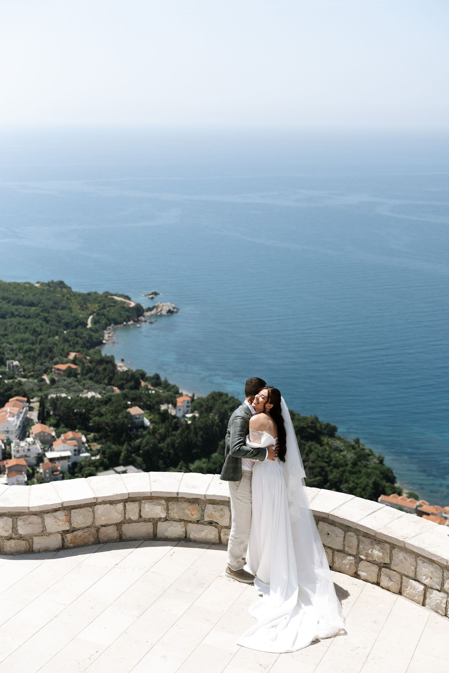 A romantic couple in wedding attire shares an intimate moment on a stone terrace overlooking the Mediterranean coastline.