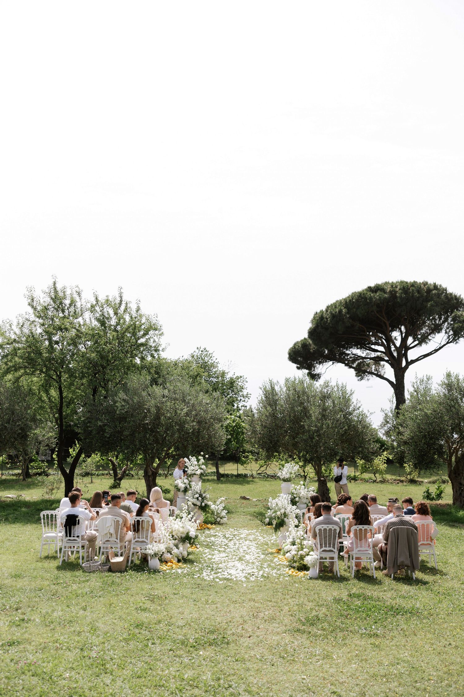 Outdoor wedding ceremony setup with white chairs arranged on a grassy lawn beneath scattered trees in a rustic setting.