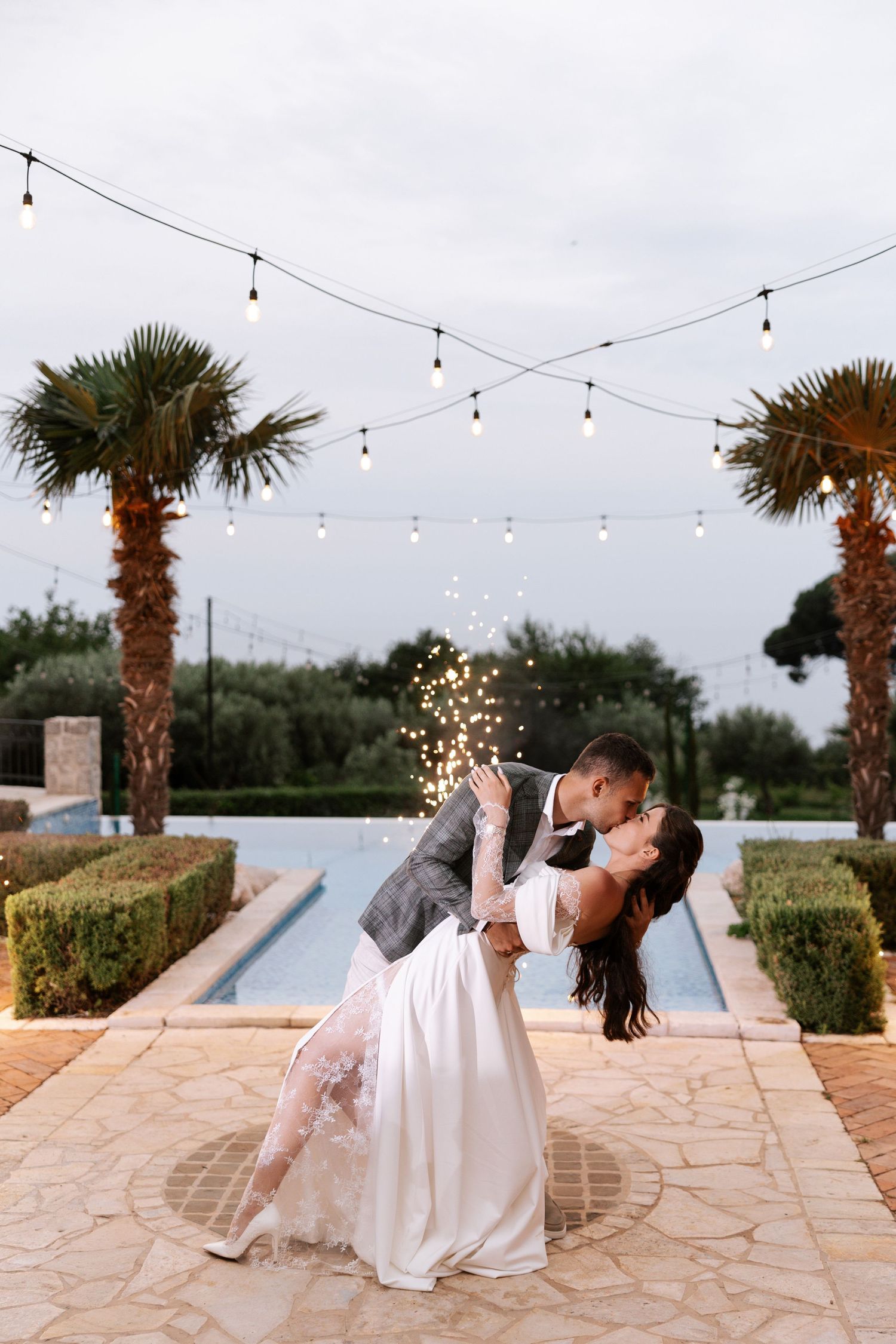 A romantic wedding couple dances and dips on a stone patio under string lights at sunset with palm trees in the background.