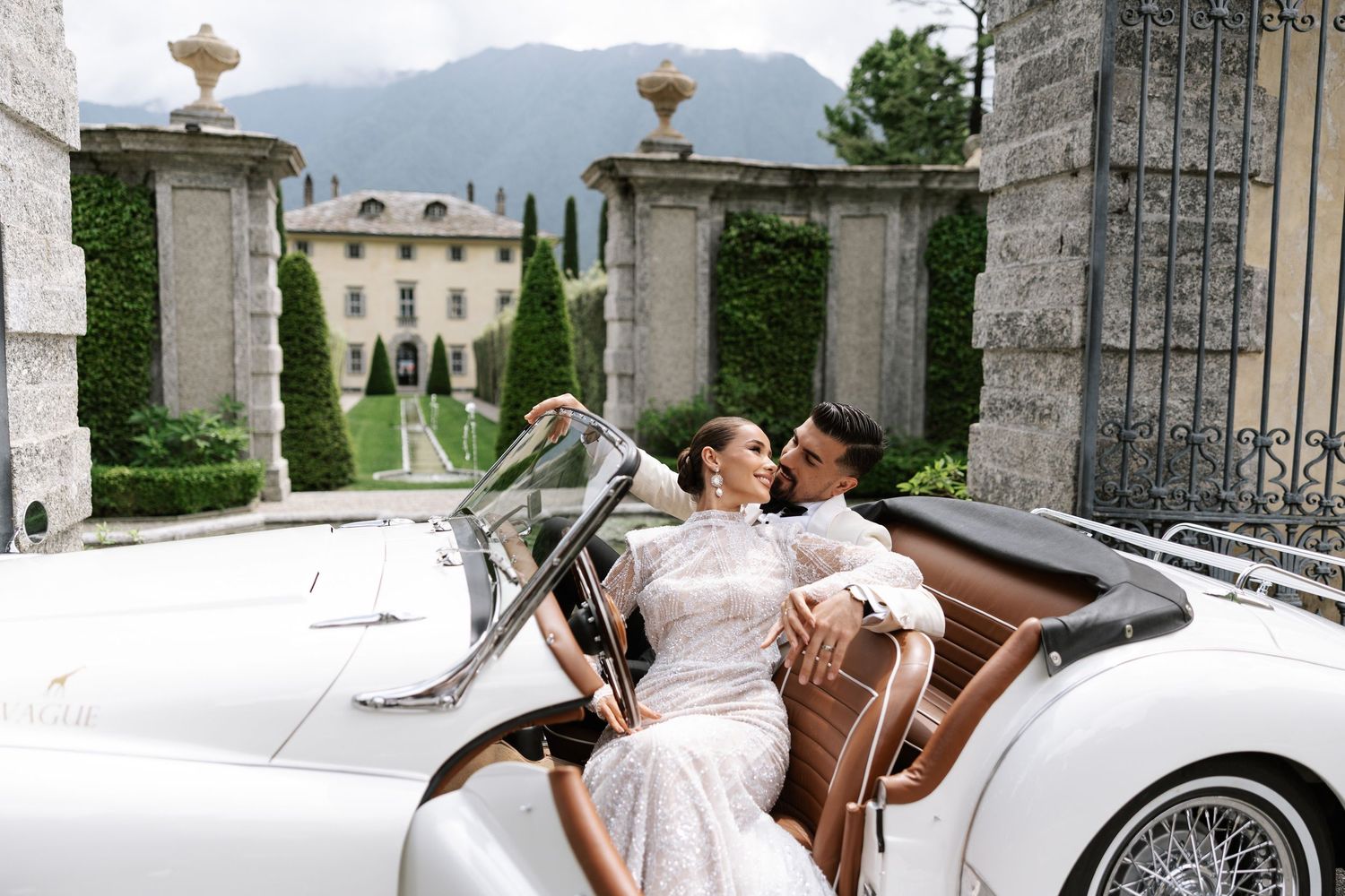 Elegant couple kissing in a vintage white convertible car outside a grand villa with mountain views.