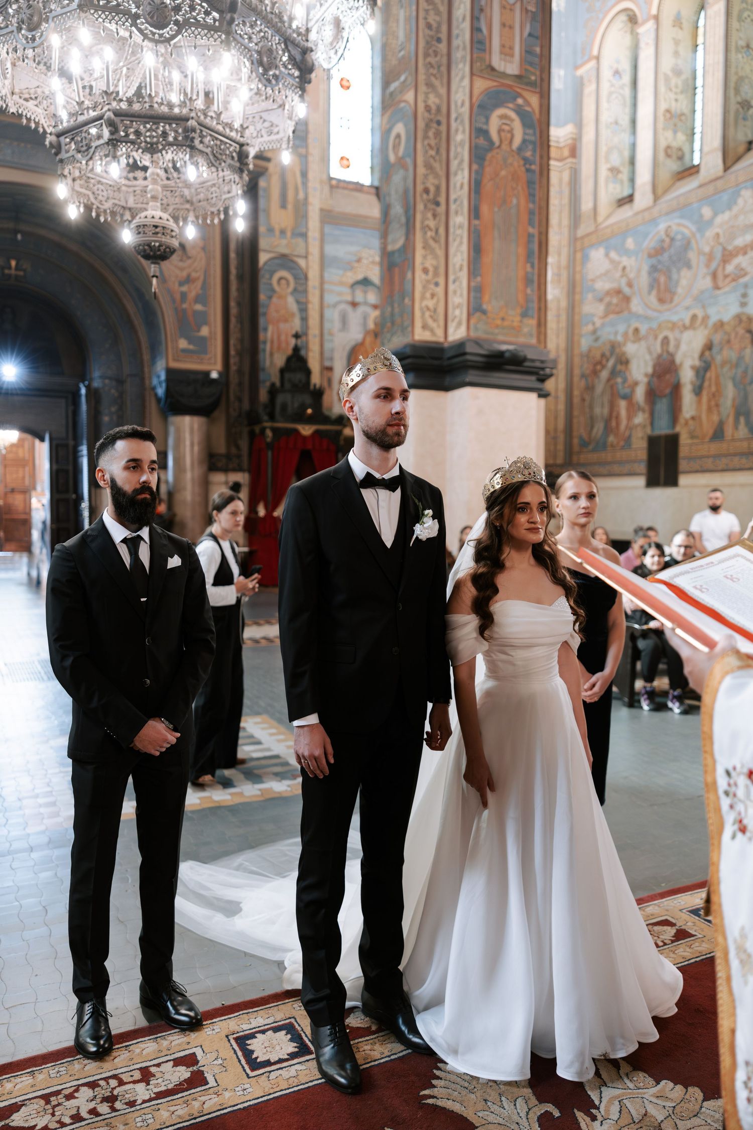 Wedding ceremony in progress beneath ornate chandeliers in decorated orthodox church.