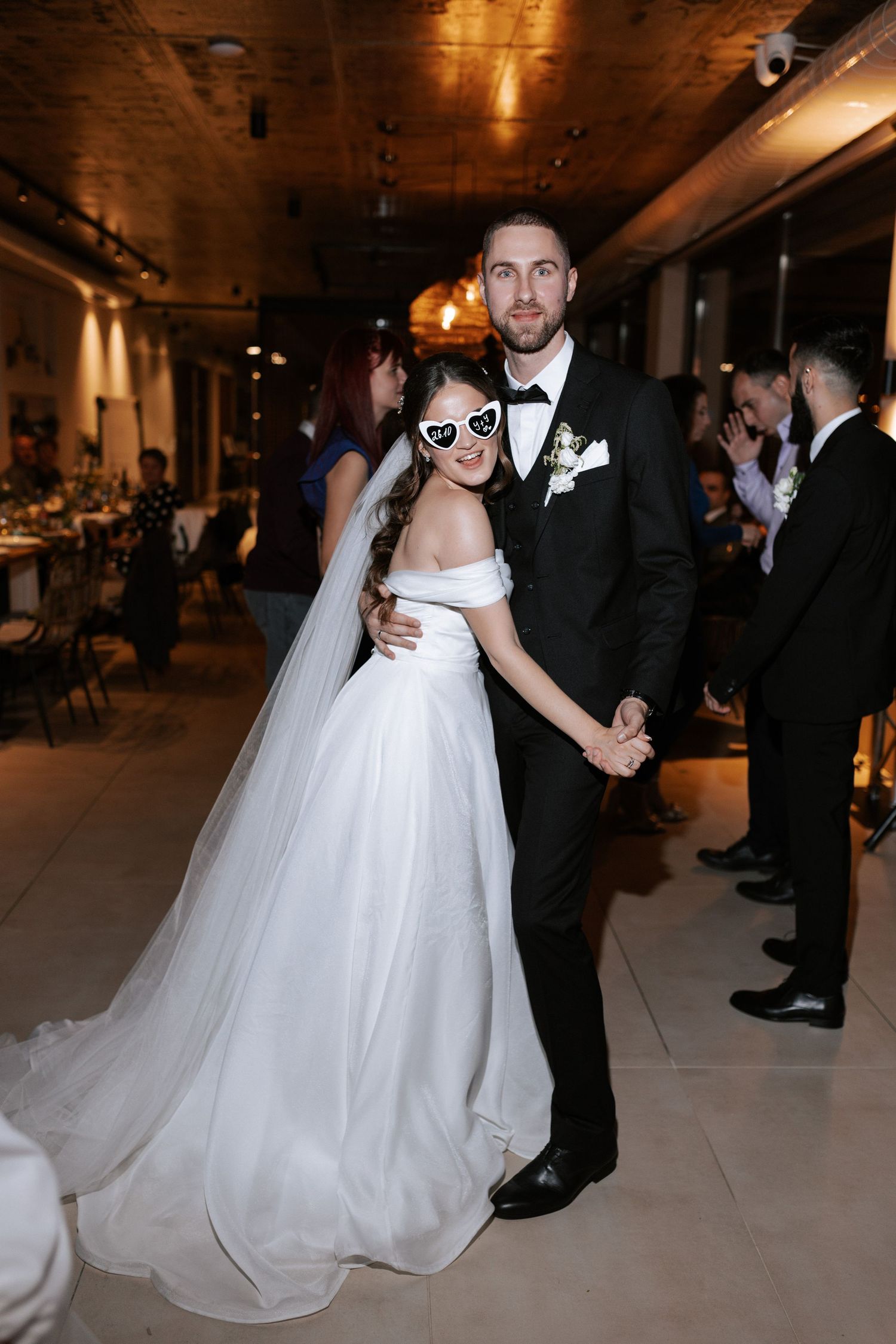 A couple poses together at their reception wearing matching heart sunglasses with their wedding attire.