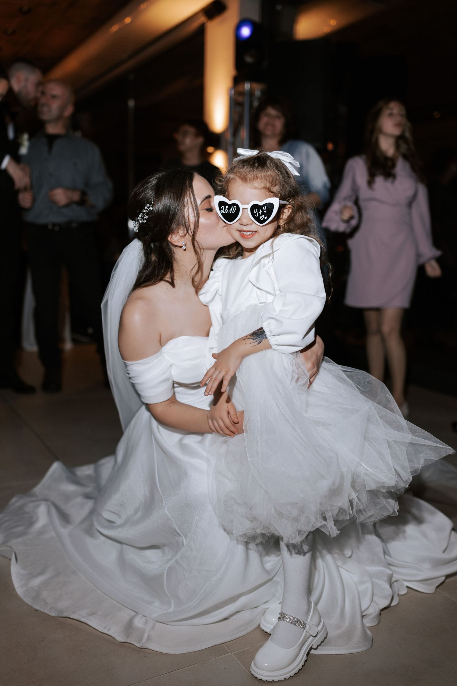 A sweet moment during a wedding reception as a bride in white dress dances with a child wearing heart-shaped sunglasses.