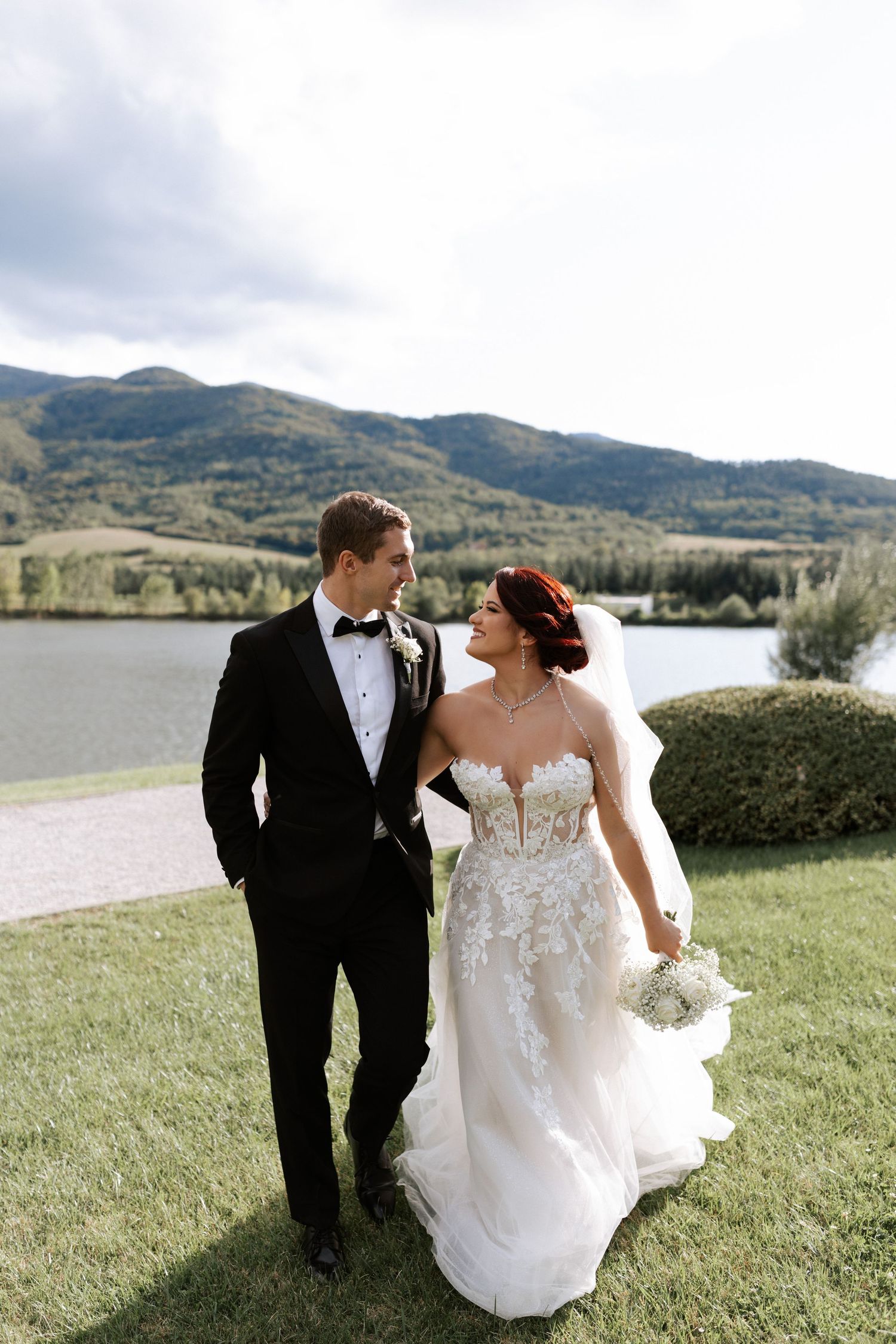 Newlyweds walk together on grass with mountain lake scenery behind them.