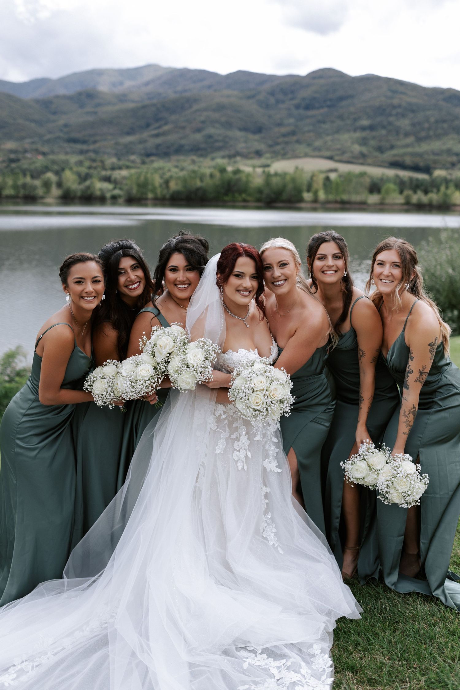 Bridal party in sage green dresses poses by a mountain lake on a cloudy day.