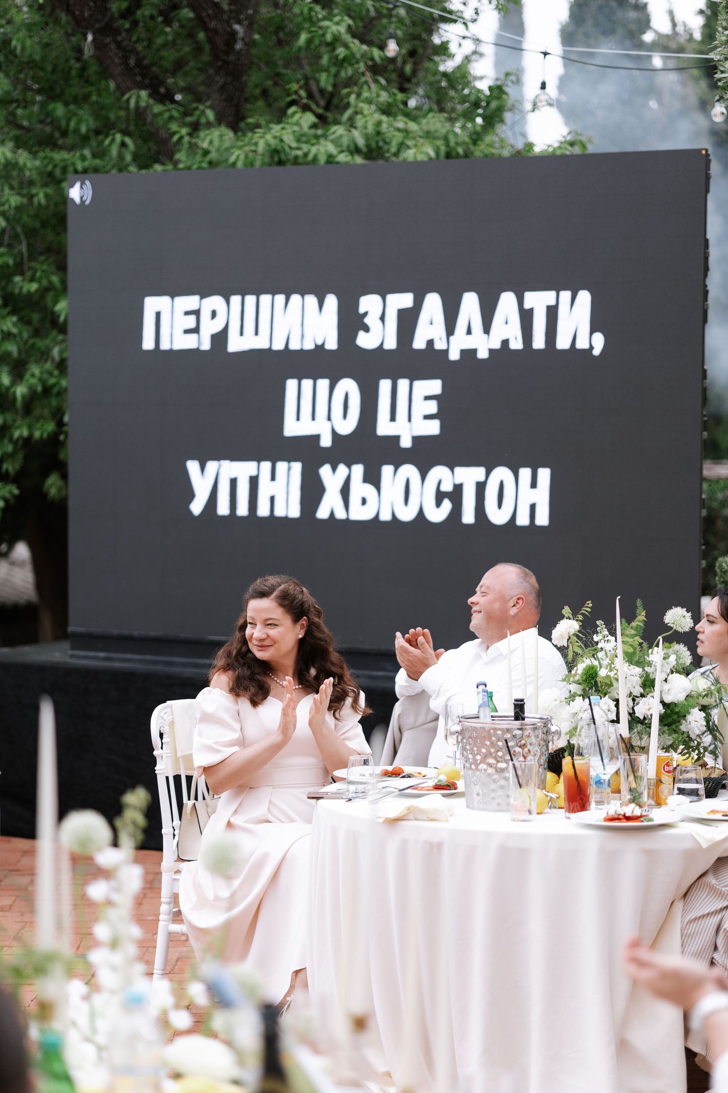 Outdoor wedding reception setup with white tables and chairs decorated with floral arrangements under trees.