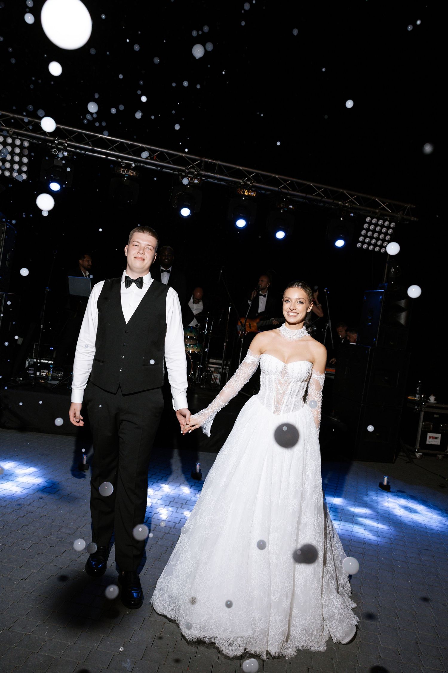 Wedding couple dancing under spotlights with falling snow effect on dark dance floor.