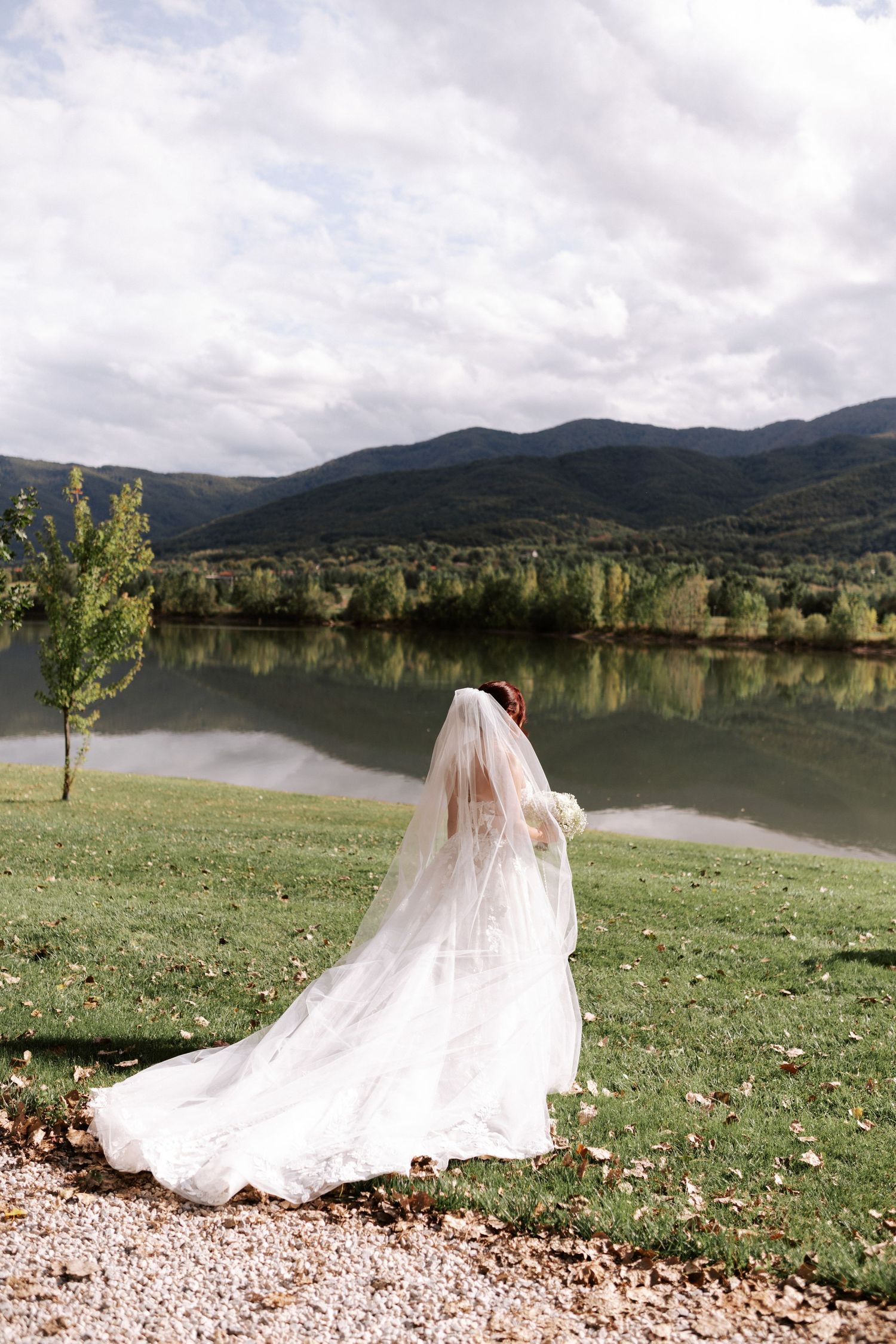 Bride's flowing white dress and veil trail behind her as she looks out over a mountain lake.