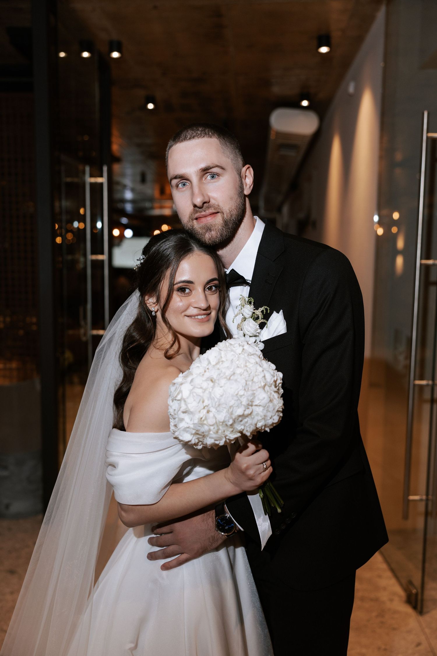 A romantic wedding portrait shows a couple dressed in formal attire with a white bouquet in an elegant indoor setting.
