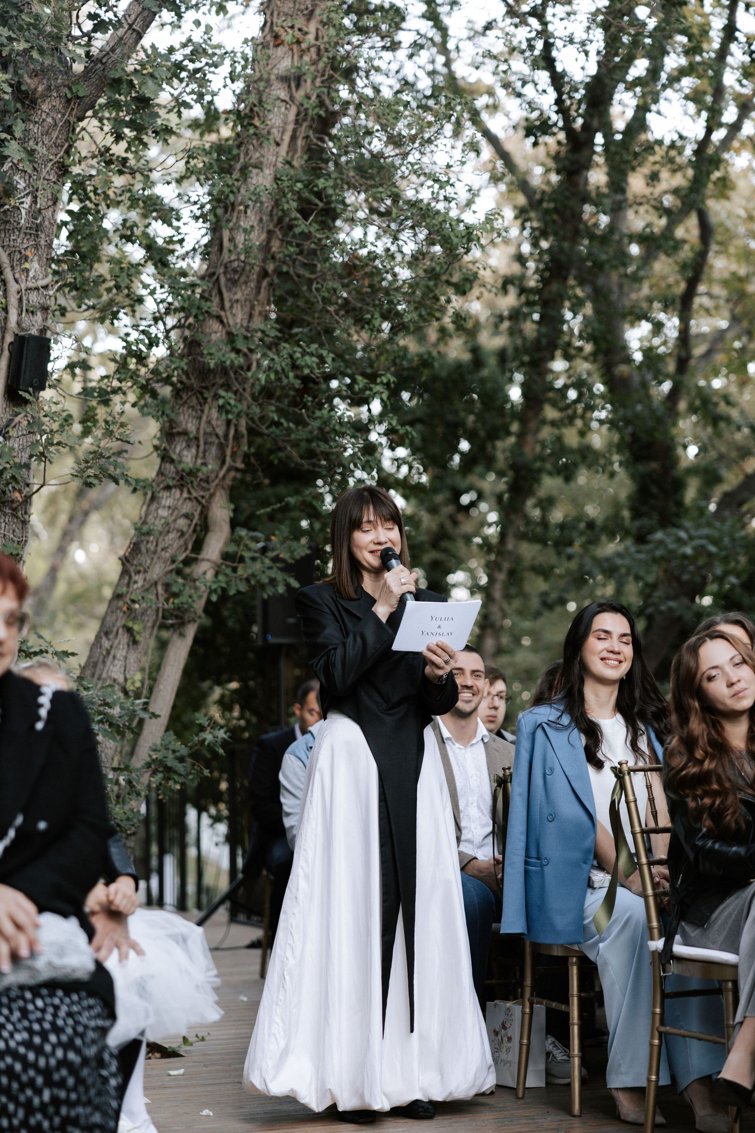 Wedding guests gather on a wooden deck surrounded by trees during an outdoor ceremony.