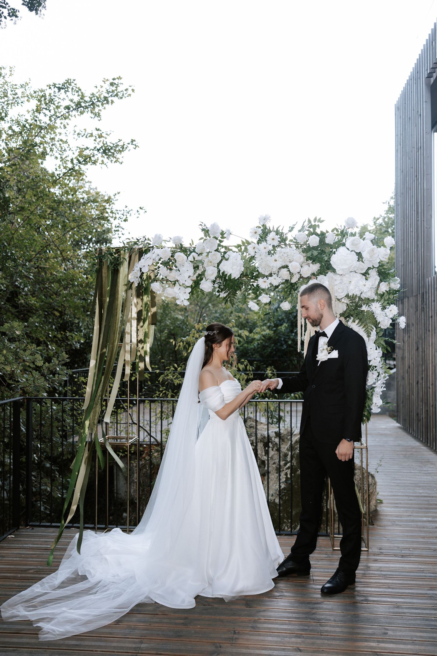 Wedding couple exchanging rings under an elegant white floral arch on an outdoor deck.