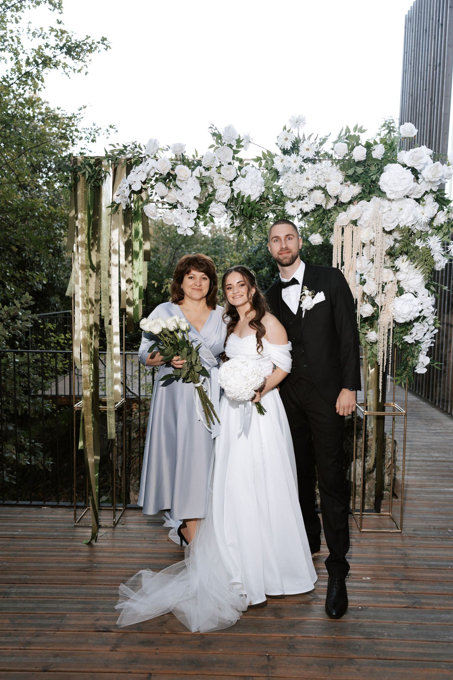 Wedding group photo under elegant white floral arch with gold accents.