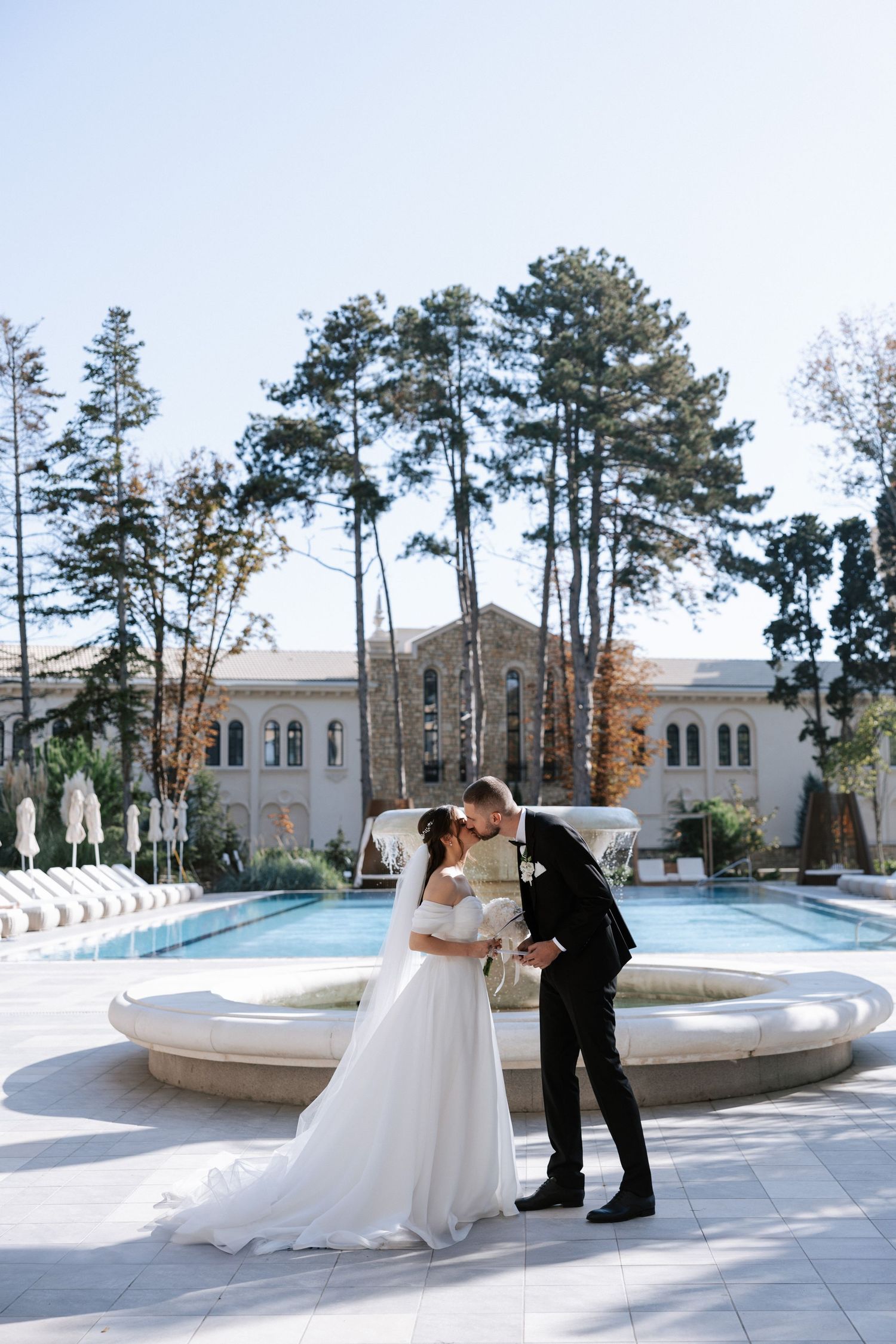 Romantic wedding couple embraces by fountain with classical architecture and pine trees in background.