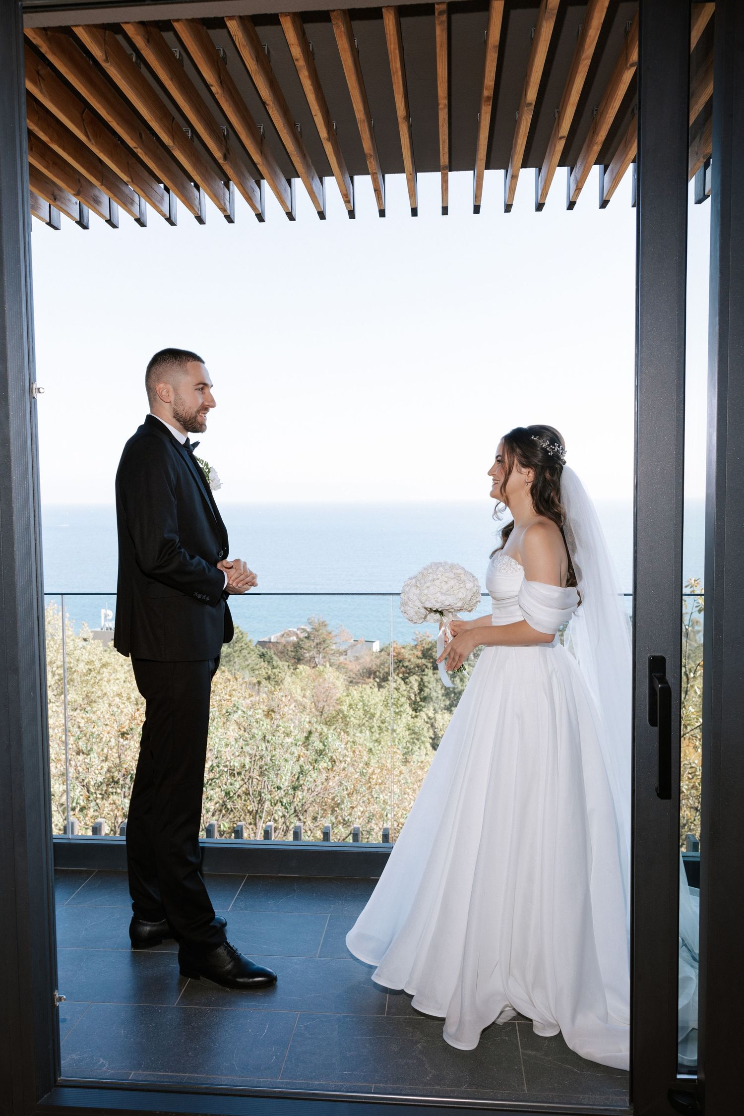 Elegant wedding couple stands beneath a modern pergola overlooking coastal scenery.