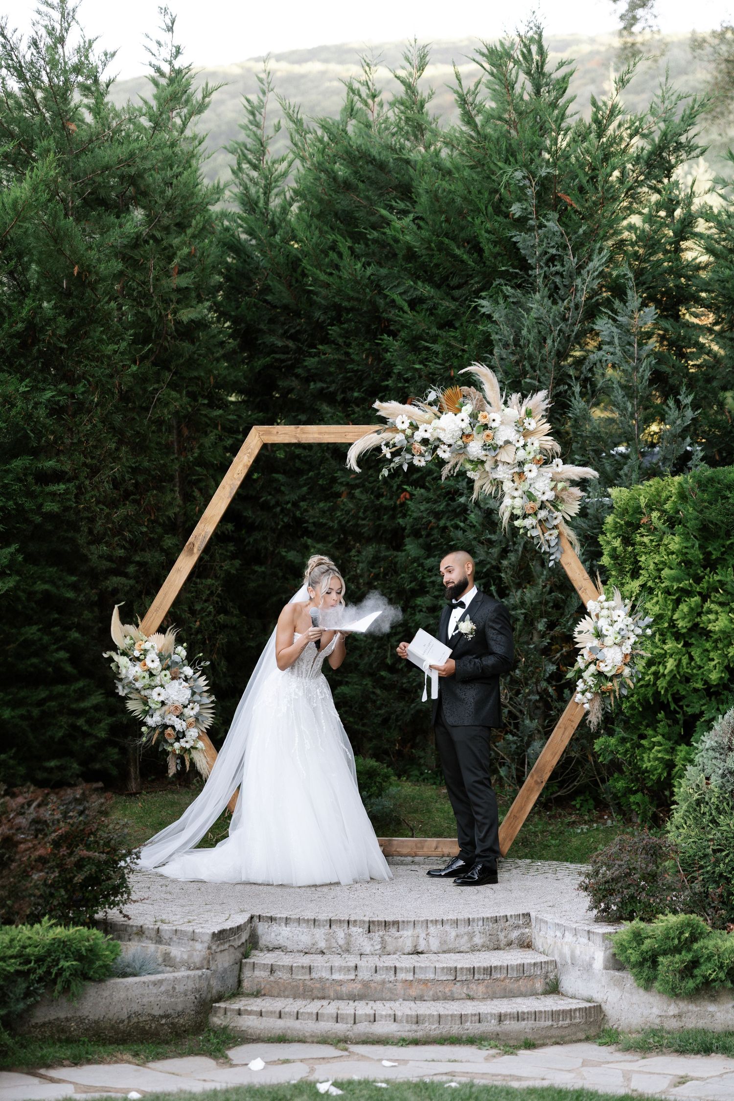 Wedding ceremony takes place at a stone patio with hexagonal arch decorated with white floral arrangements and greenery.