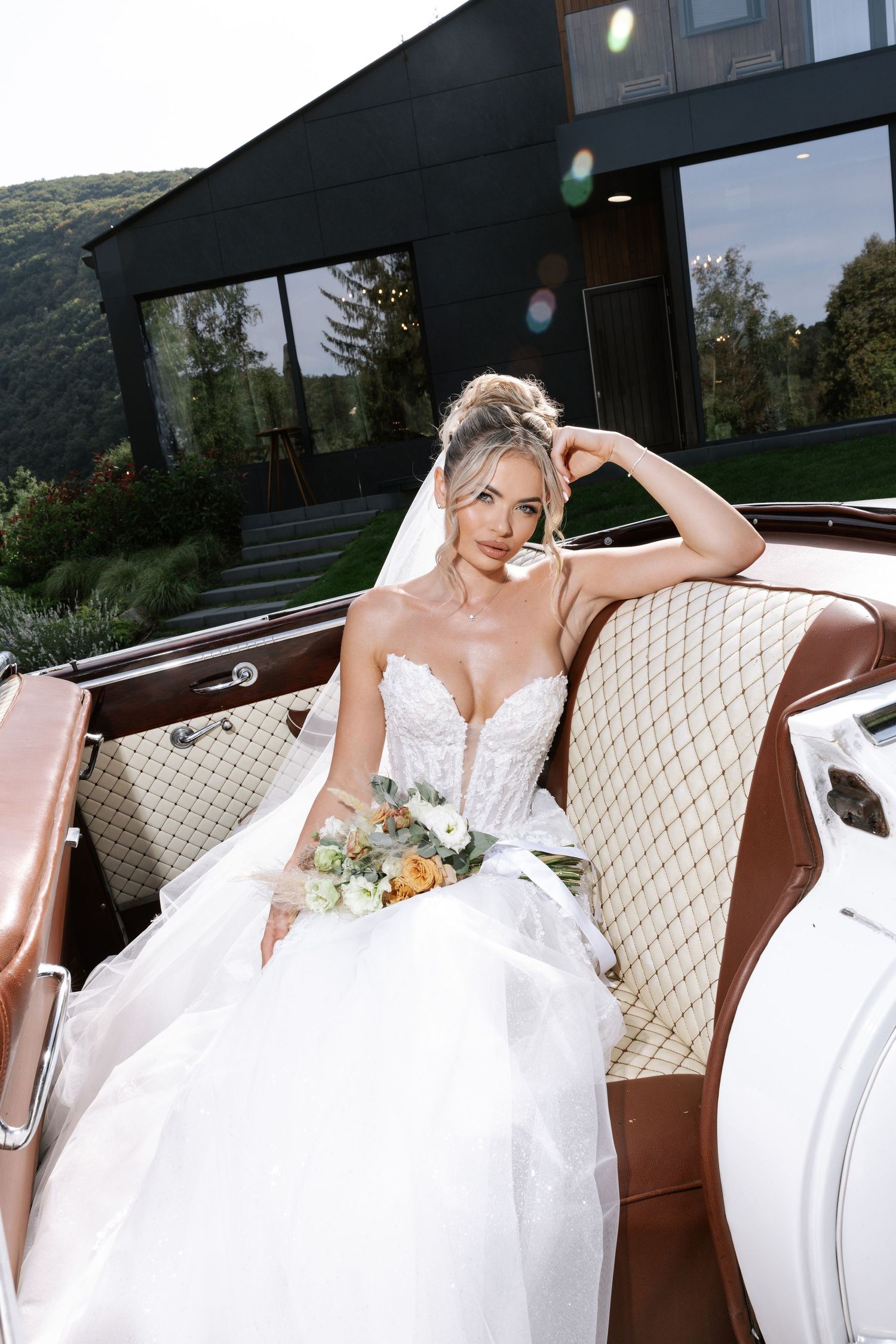 A bride in a white wedding gown poses elegantly on cream-colored seating in a vintage vehicle interior.