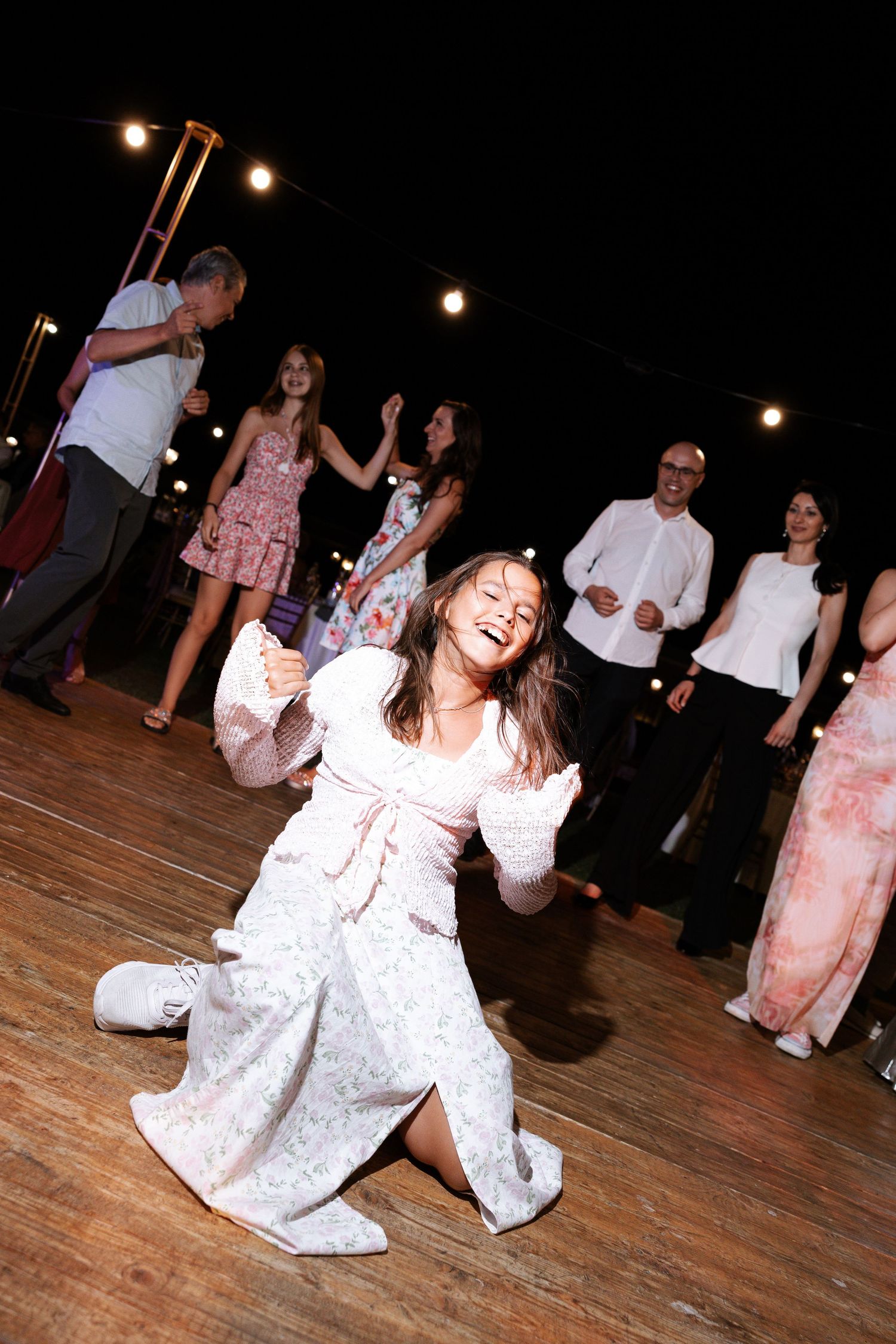People dancing together on a wooden dance floor at a party with string lights overhead.