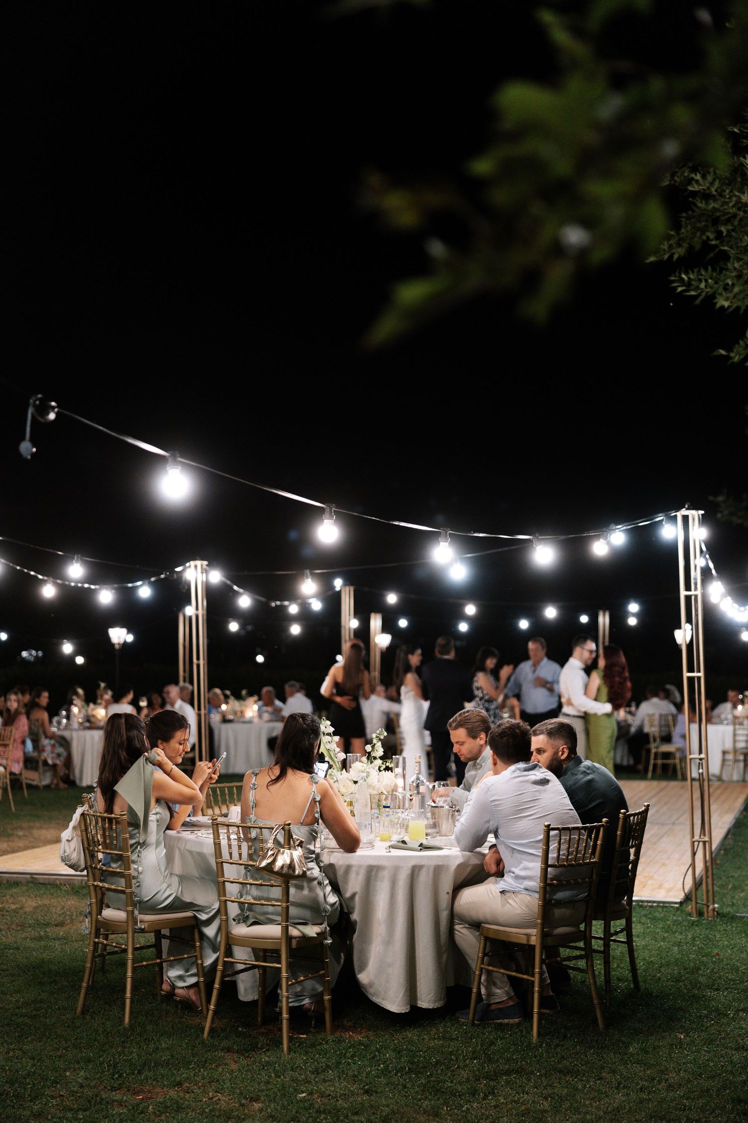 String lights illuminate an elegant outdoor wedding reception with guests seated at round tables on a grassy lawn at night.