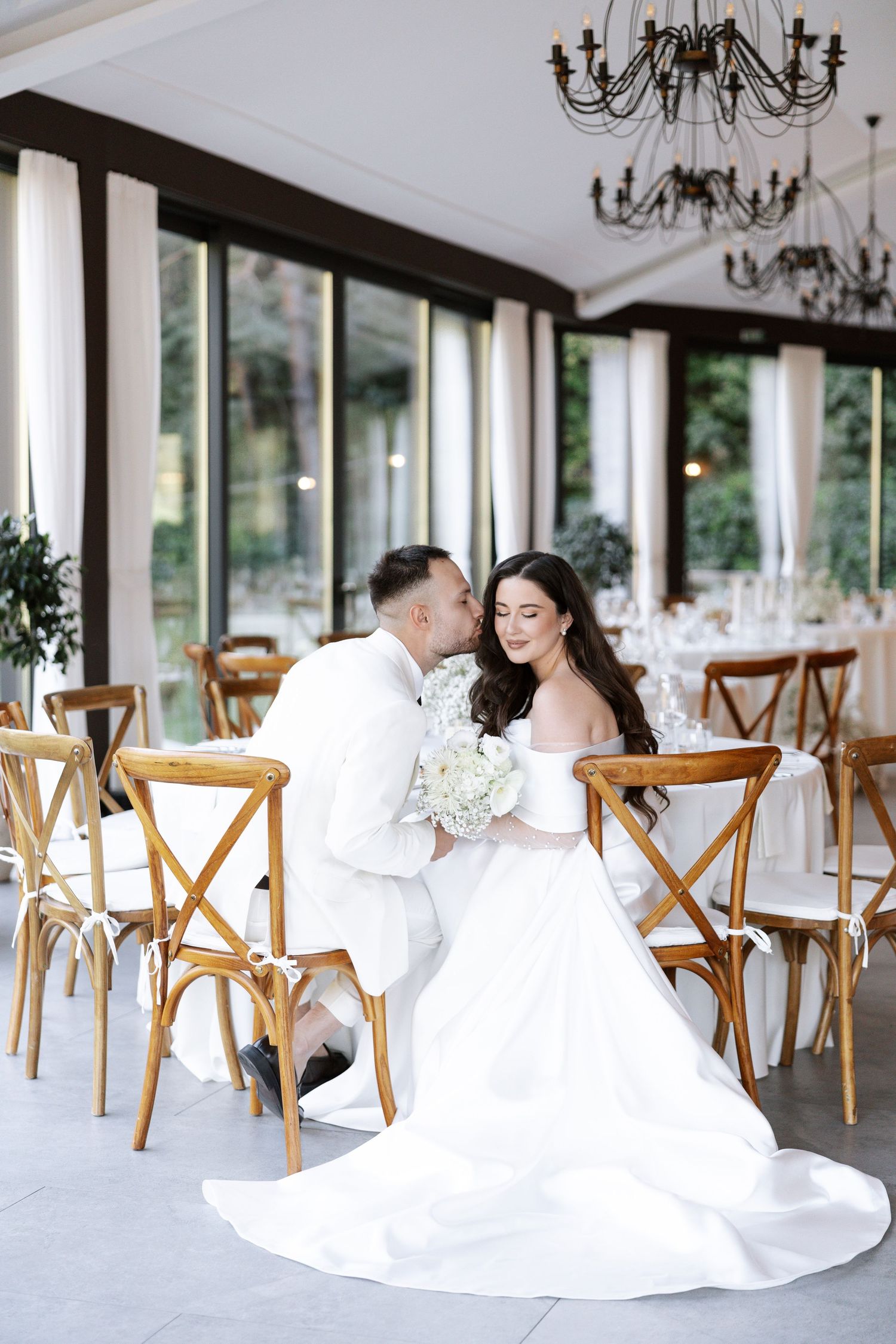 Elegant wedding reception venue with wooden cross-back chairs and crystal chandeliers set in bright white room.