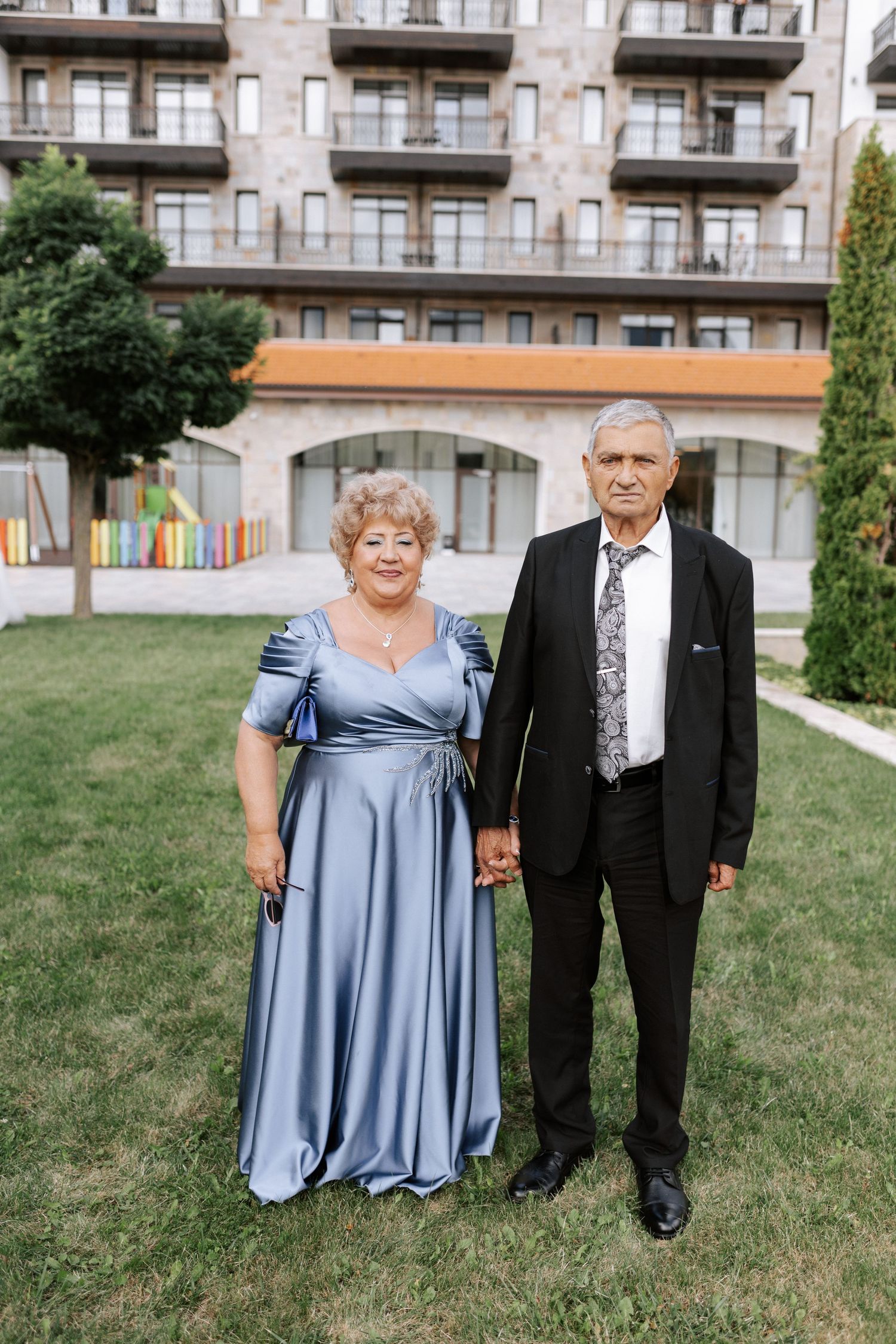 Couple in formal attire poses on lawn in front of modern apartment building.
