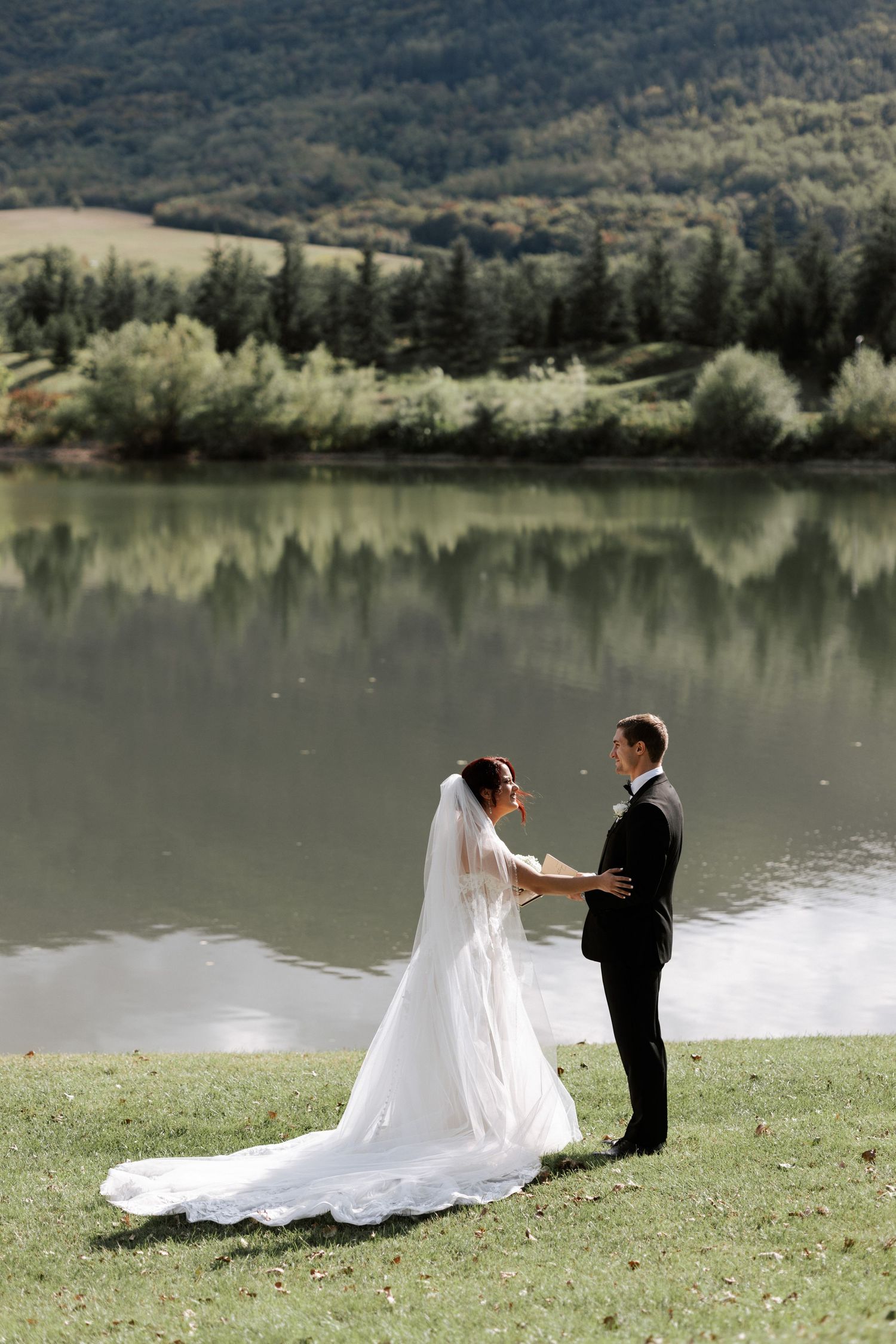 A couple exchanges wedding vows on a grassy shore with a mirror-like lake reflection behind them.