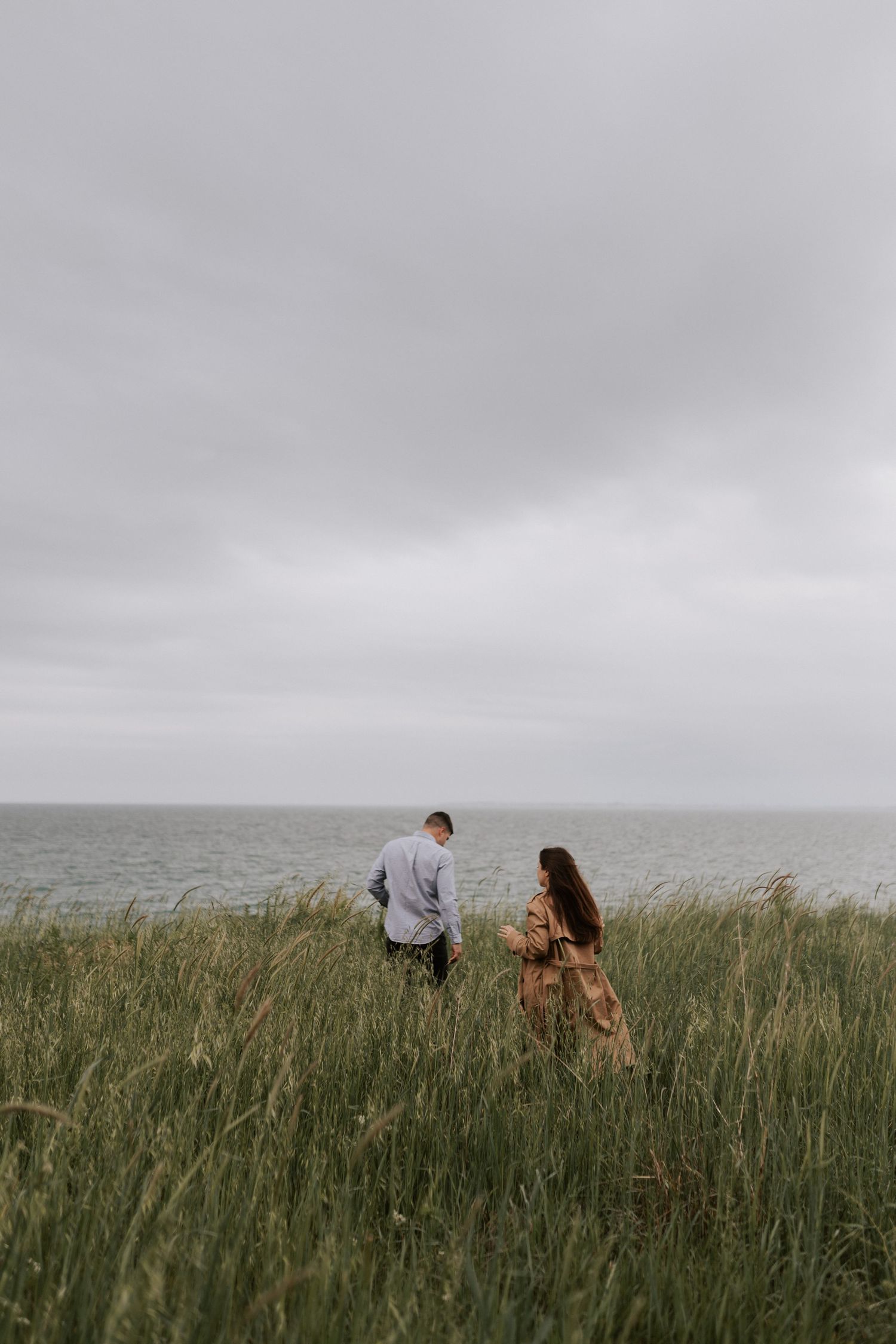 Couple walking through tall coastal grass overlooking the ocean on an overcast day.