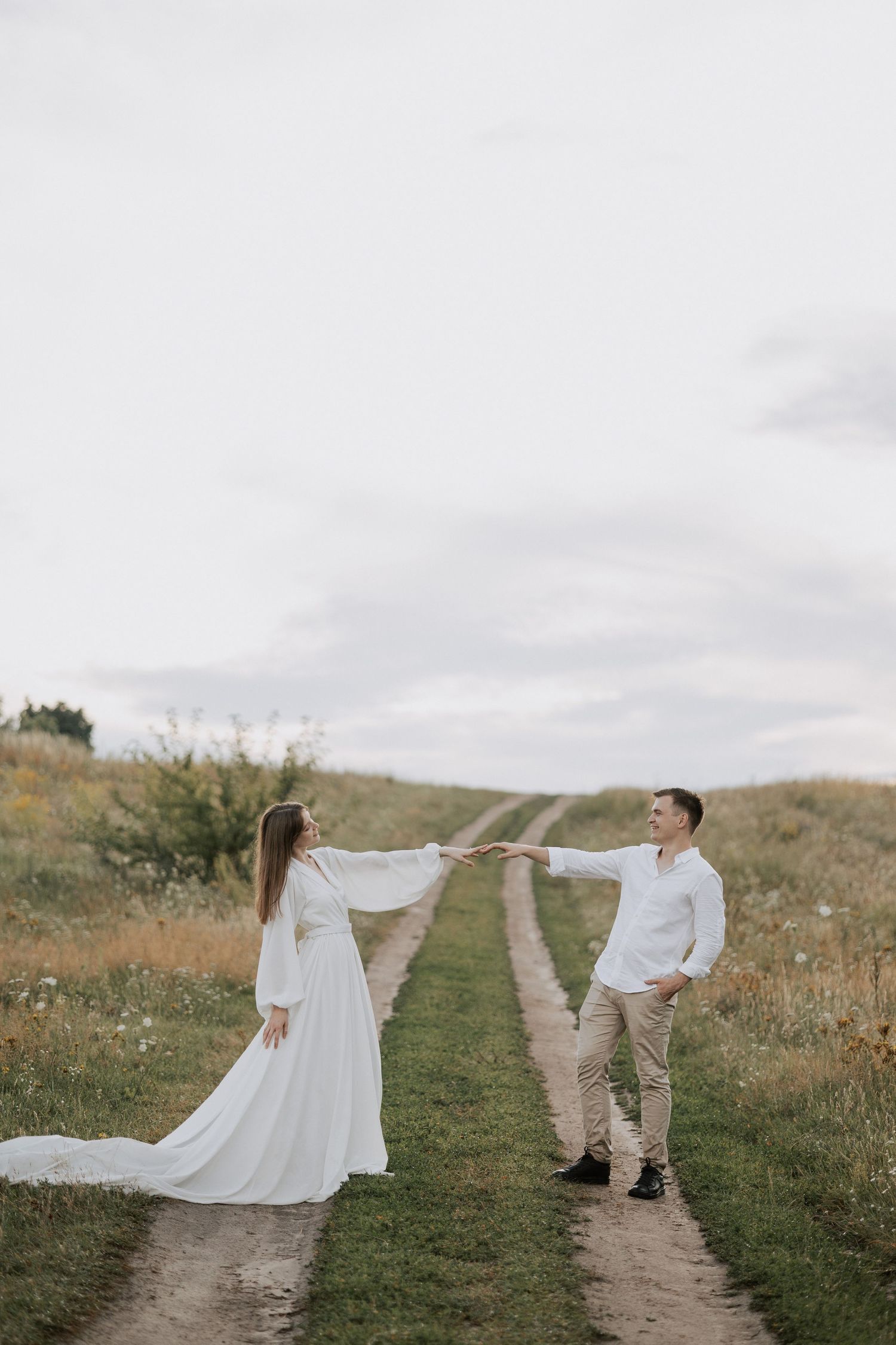 Couple in white outfits holding hands on rustic path through meadow.