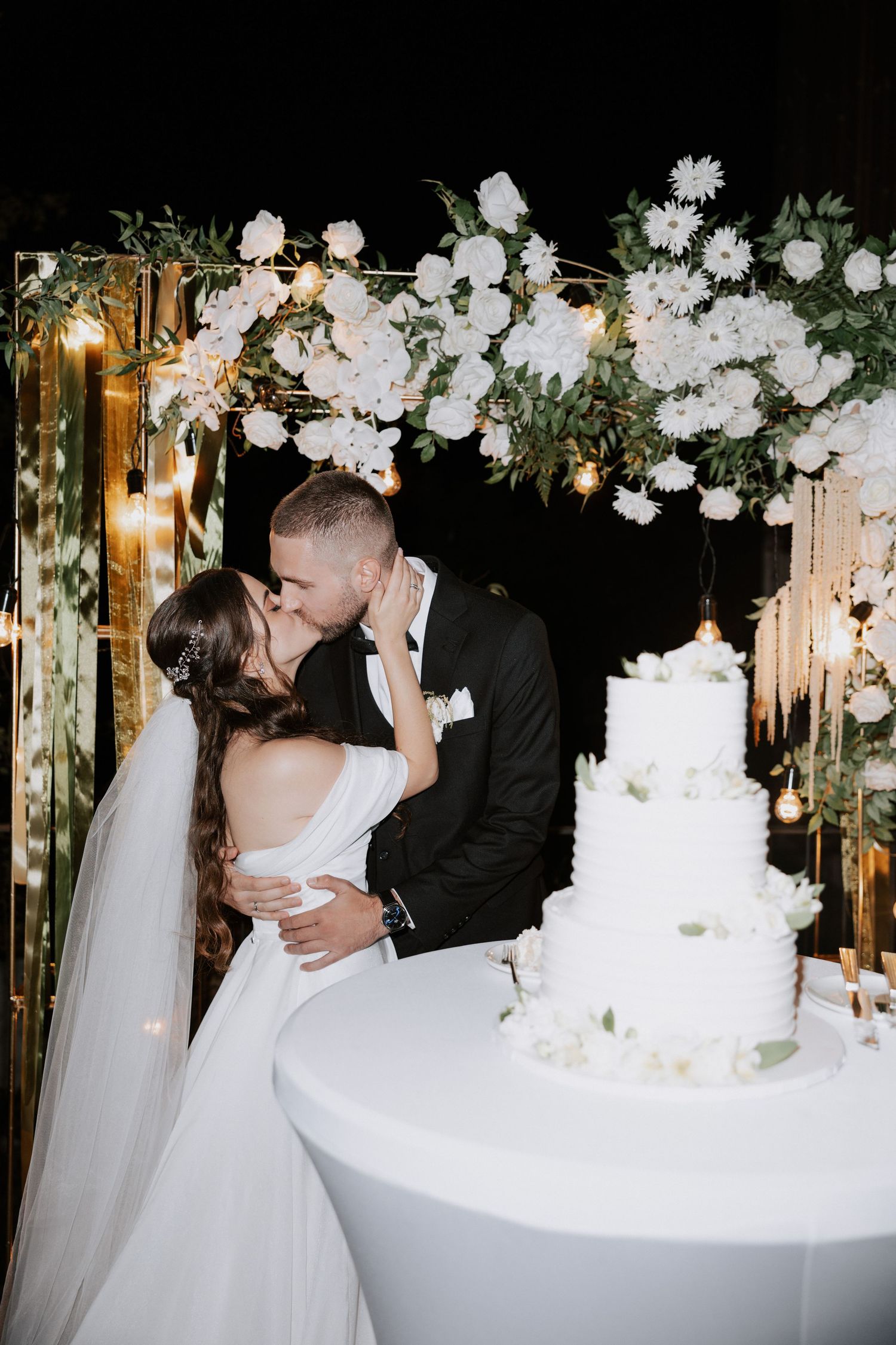Newlyweds share a romantic kiss by their wedding cake under beautiful white floral arrangements.