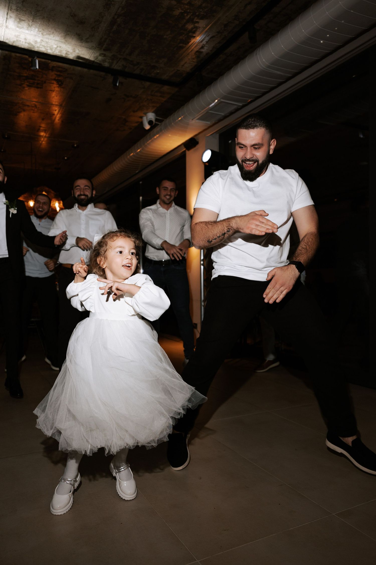 A young girl in a white dress dances energetically next to a person in a white t-shirt during a celebration.