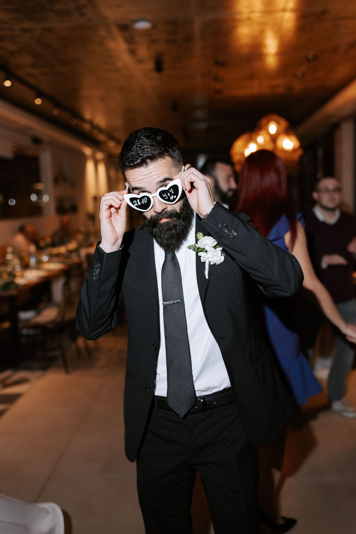 A guest in formal attire adjusts heart-shaped sunglasses at a wedding reception.