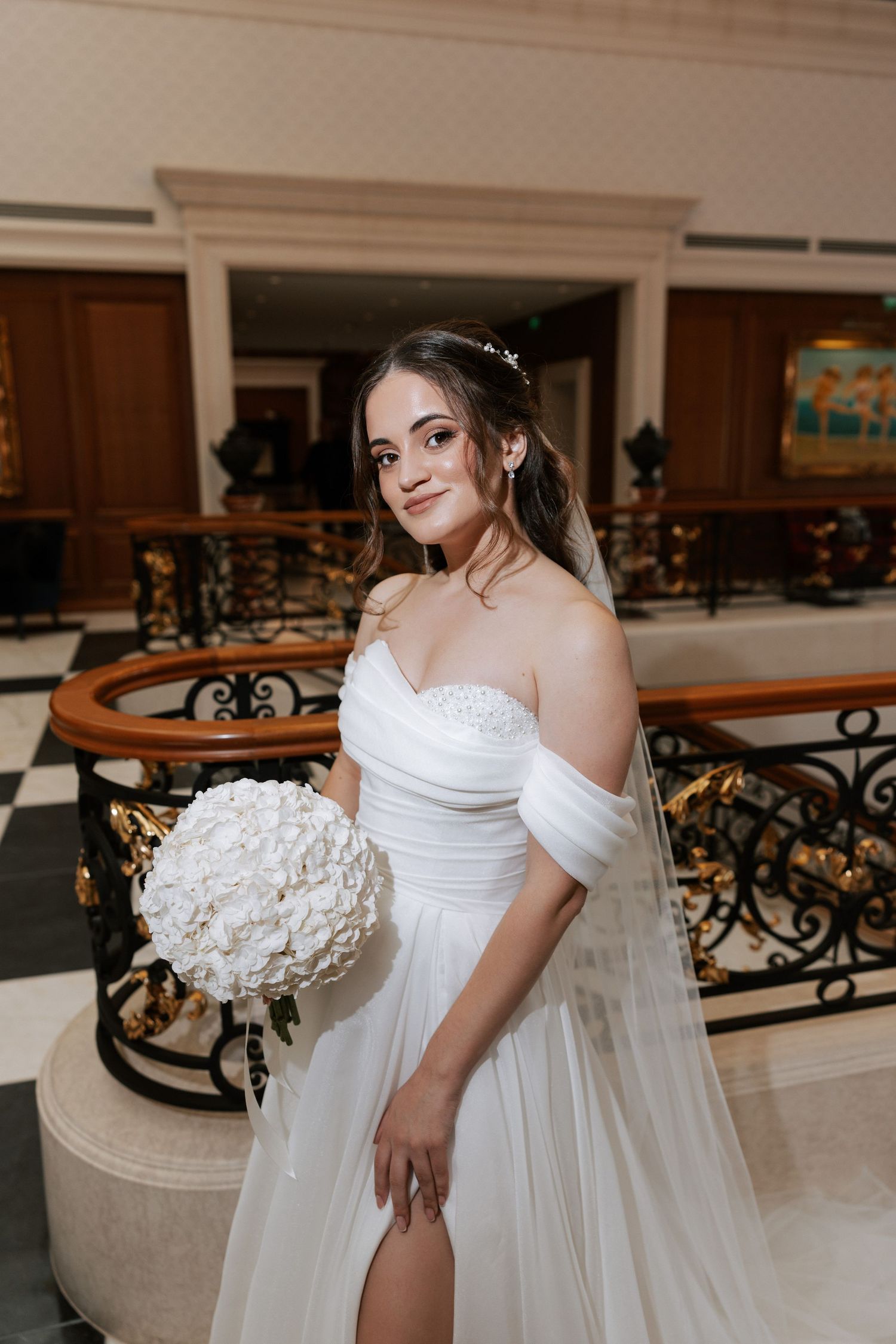 A bride in an off-shoulder white wedding dress holds a white floral bouquet while posing in an elegant venue.