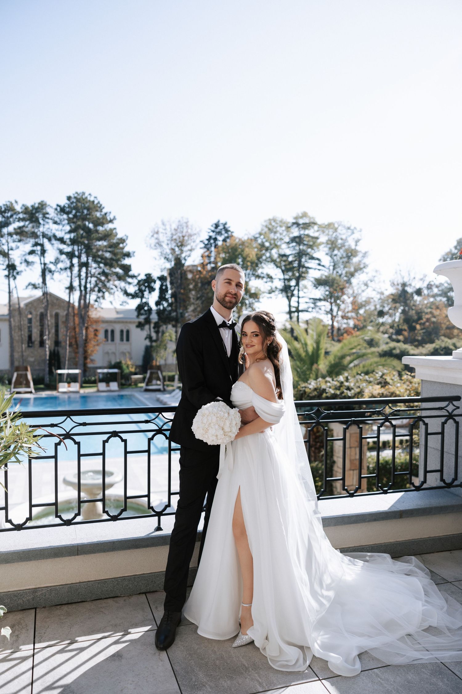 Wedding couple poses on balcony overlooking pool area with tall pine trees in background.