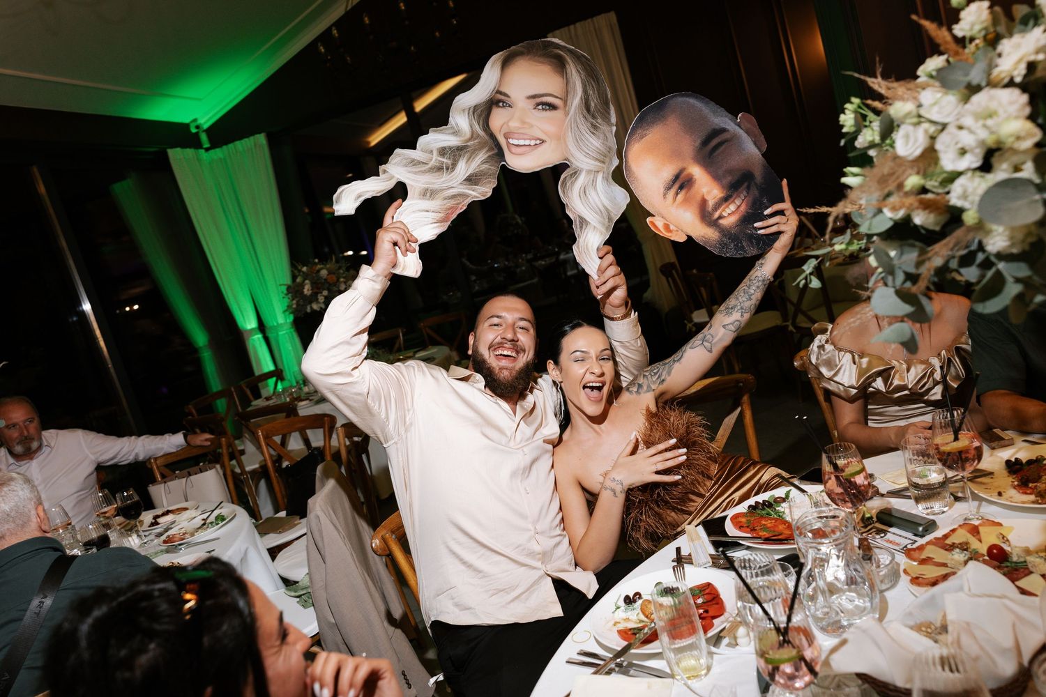 Wedding guests celebrate and dance during a lively reception with green lighting and white floral decorations.